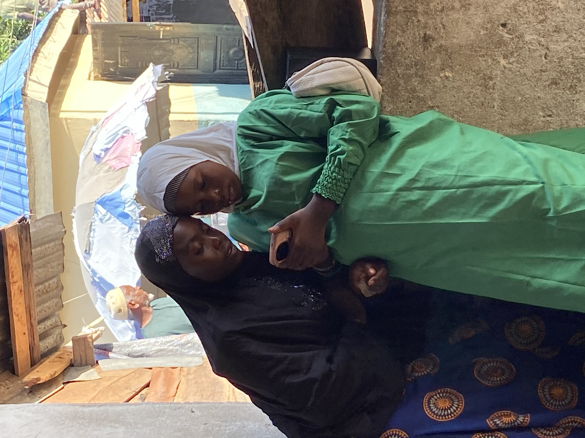 Two young women are together indoors. One woman is in a green dress and white hijab, and the other woman is in a black hijab and a dark patterned dress. They are holding hands. An elderly man is lying on a bed in the background. The setting appears to be a modest dwelling. The location is Mararaba, Nigeria.