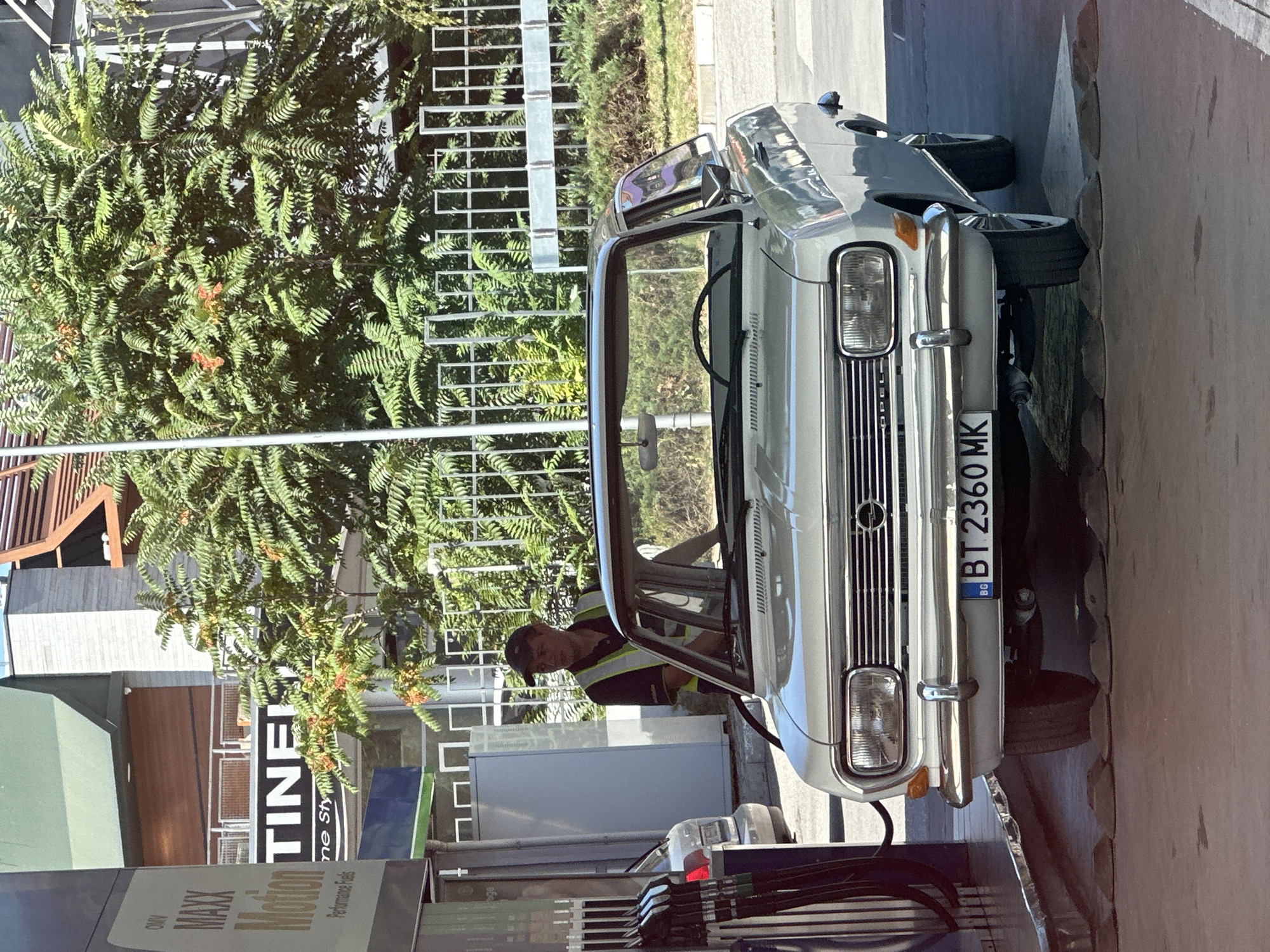 A vintage white Opel Kadett is parked at a gas station in Veliko Tarnovo, Bulgaria. A male attendant wearing a high-visibility vest and a dark cap is standing by the car, seemingly in the process of refueling it. The gas station appears to be part of a larger commercial complex, with signage for "OMV MAXX Motion Performance Fuels" visible. In the background, there are lush green trees and a modern building with a slatted roof. The license plate of the car reads "BT 2360 MK." The scene is well-lit, suggesting it is daytime.