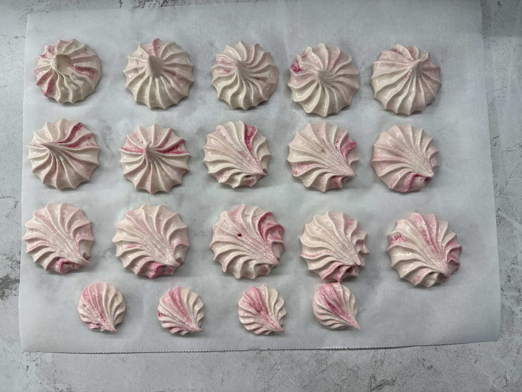 The image shows a top-down view of twenty-one meringues arranged on a sheet of white parchment paper. The meringues are white with streaks of pink, suggesting they are strawberry or raspberry flavored. They are arranged in four rows: the top row has four meringues, the second row has six, the third row has six, and the bottom row has five. The background is a speckled gray surface, possibly a countertop or table. There is no visible indication of the location being Lyubertsy, Russia, or any other specific geographical place. The image is a still life of baked goods.
