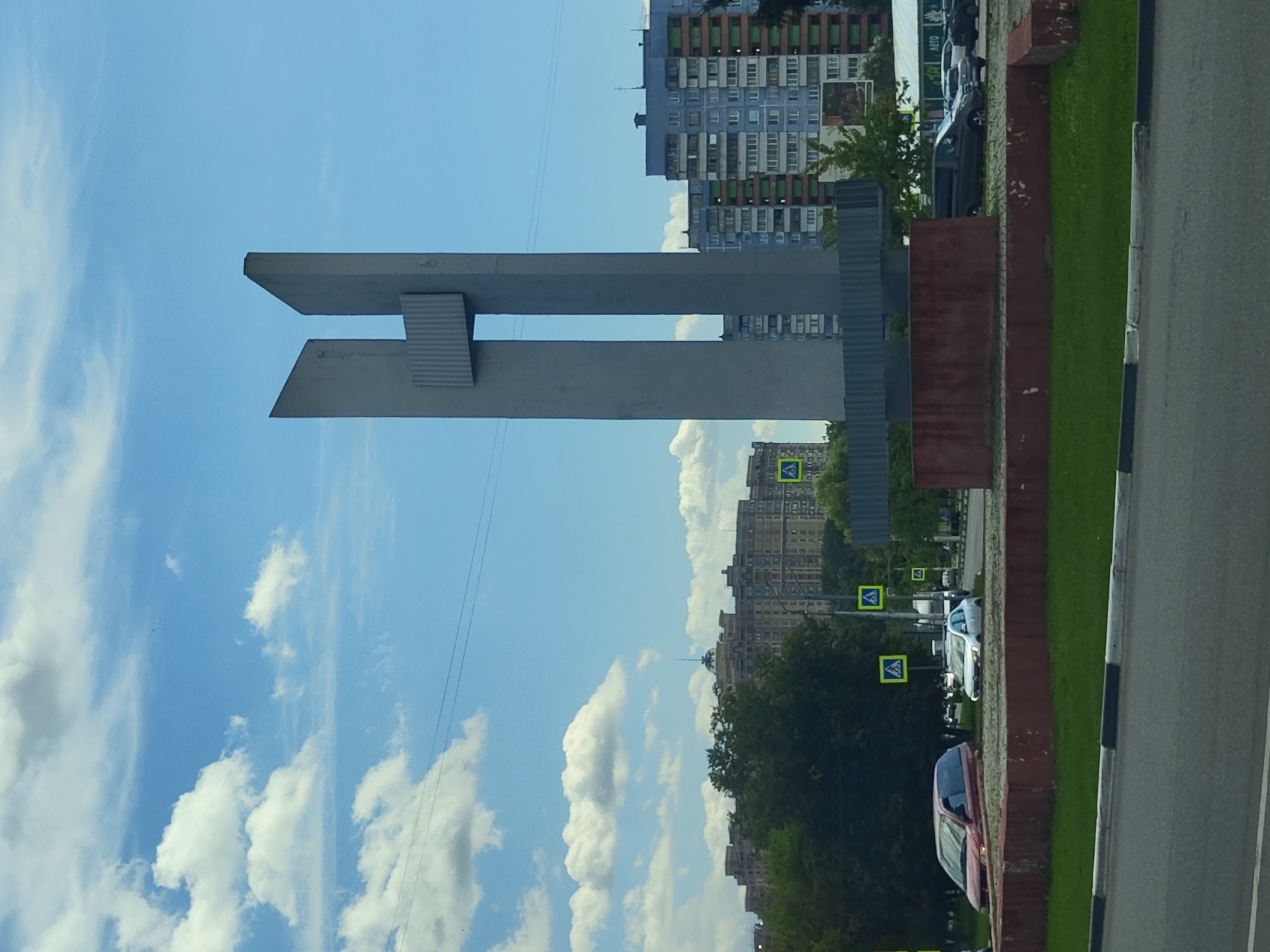 The image shows a modern sculpture in the city of Ramenskoye, Russia, situated in an urban environment with residential buildings, trees, and a road. The sculpture is tall and abstract, made of concrete, with a distinctive H-like shape. It stands on a raised, reddish-brown concrete base surrounded by green grass.
In the foreground, a road with a white and black curb is visible. To the left of the sculpture, a red minivan is parked on the grass area. To the right, a silver sedan is also parked, and further to the right, a dark-colored van is parked next to other vehicles.
Several pedestrian crossing signs, indicated by yellow triangles with a white pedestrian symbol, are placed around the area. In the background, large residential apartment buildings are prominent. On the left, a long, multi-story building with a rounded top and a spire is visible behind trees. To the right, several multi-story apartment blocks with varied facade colors, including blue, white, and orange, dominate the skyline.
The sky is a bright blue with scattered white cumulus clouds. Power lines are visible stretching across the upper portion of the image. The overall scene suggests a clear, sunny day in a developed urban area.
