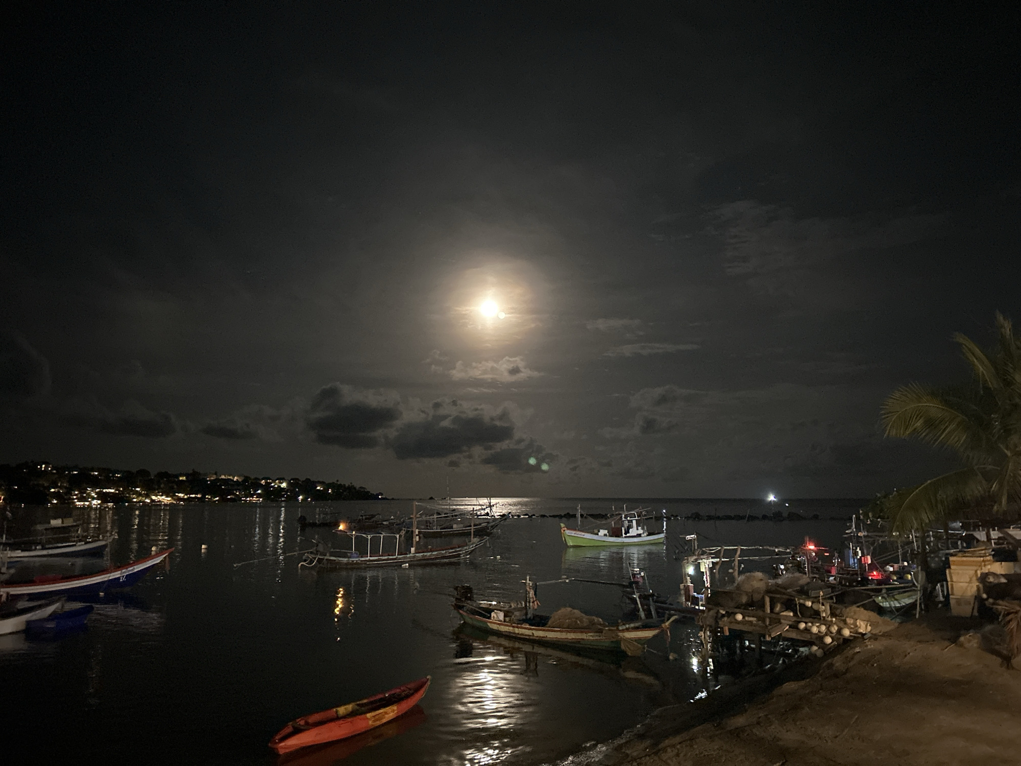 The image depicts a nighttime scene at Baan Chaweng Noi, Thailand. In the foreground is a body of water with several boats of varying sizes and conditions, some docked close to shore, others anchored further out. A small, red kayak is visible near the shoreline in the lower left quadrant. Reflected moonlight creates a bright path across the water. The sky is overcast, with a visible full moon shining through the clouds. Across the water, lights suggest the presence of buildings and settlements along the coast. A palm tree is present on the right side of the image. The scene is generally dark, illuminated primarily by the moon and artificial lights. The shore is made of dark earth.