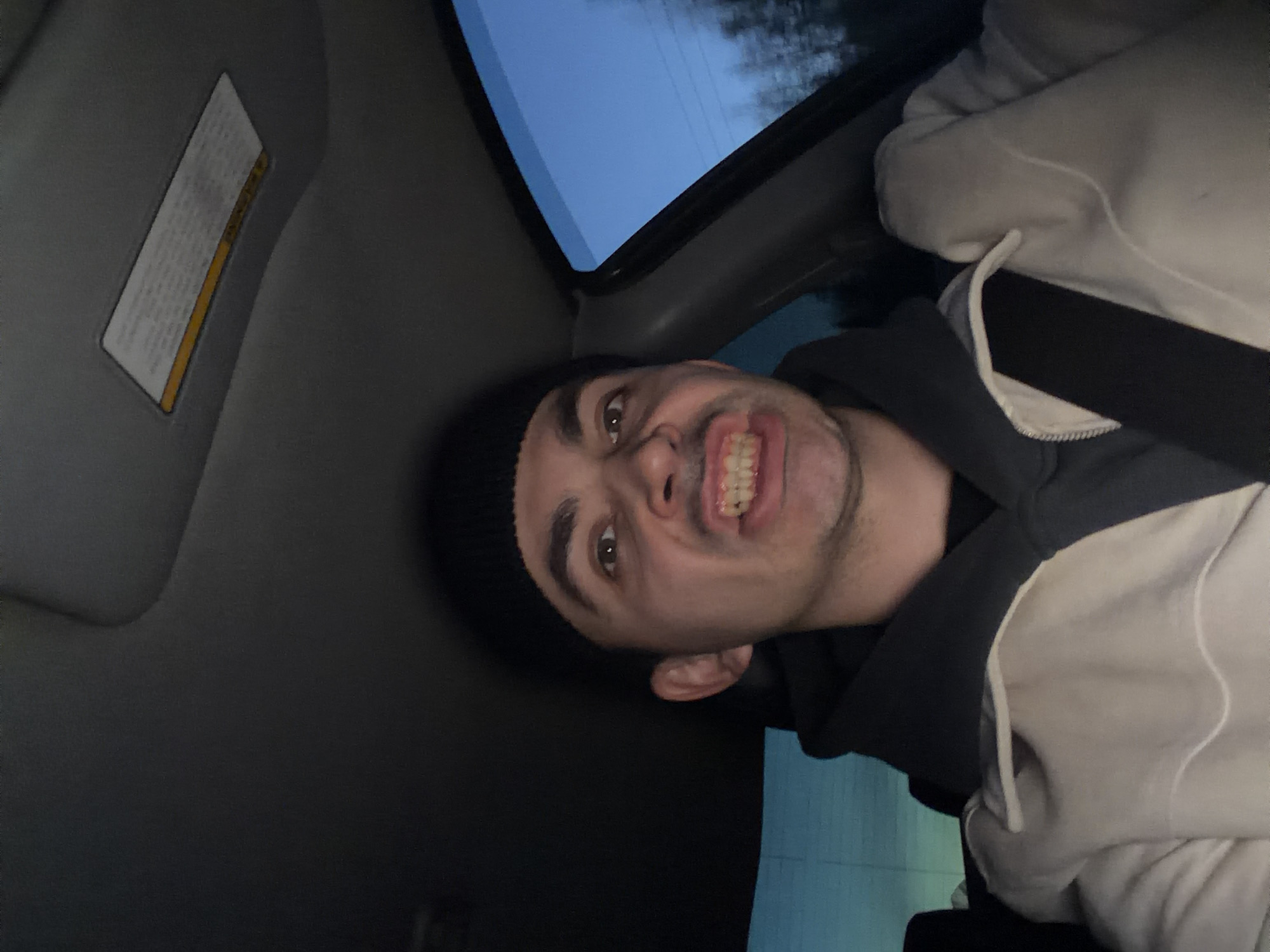 A man wearing a black beanie and a cream-colored hooded sweatshirt with a black zip-up underneath is in the driver's seat of a car. He is wearing a seatbelt. The interior of the car is visible, including the sun visor with a warning label. The sky is visible through the car window. The man is making a face, with his teeth bared. The scene appears to be taken during the day. There is no evidence to suggest that the image was taken in Shaberdino, Russia.