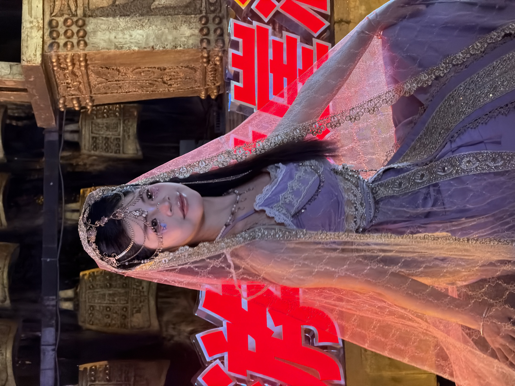 A young woman in a purple dress and veil is seated in front of a backdrop of large, ornate bells. The bells appear to be made of metal and are decorated with intricate carvings. In the foreground, there are large, bright red Chinese characters. The woman is wearing a jeweled headdress and has glitter on her face. Her dress is embellished with silver trim. The overall impression is of a cultural performance or festival. The scene is located in Yuzhong District, China.