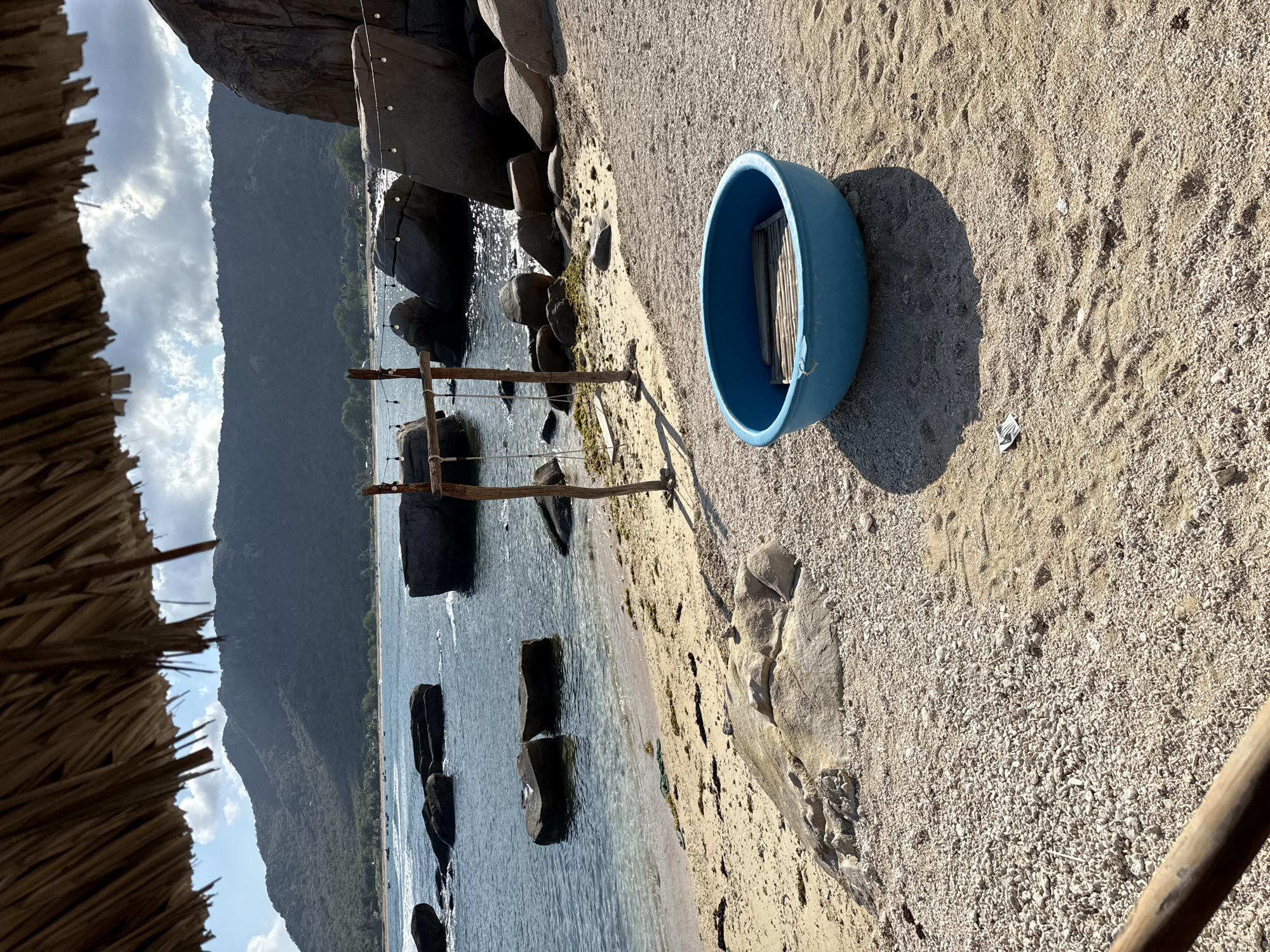 The image shows a beach scene in Vinh Hai, Vietnam. In the foreground, a bright blue, round plastic boat rests on the sandy beach. A wooden frame lies inside the boat. The beach consists of sand and small pebbles. Nearby, large rocks border the water's edge. In the middle ground, a wooden structure with strung lights is visible near the shoreline. The water is clear, reflecting the sky. In the background, a large, forested mountain rises against a cloudy sky. A thatched roof overhangs the left side of the image.