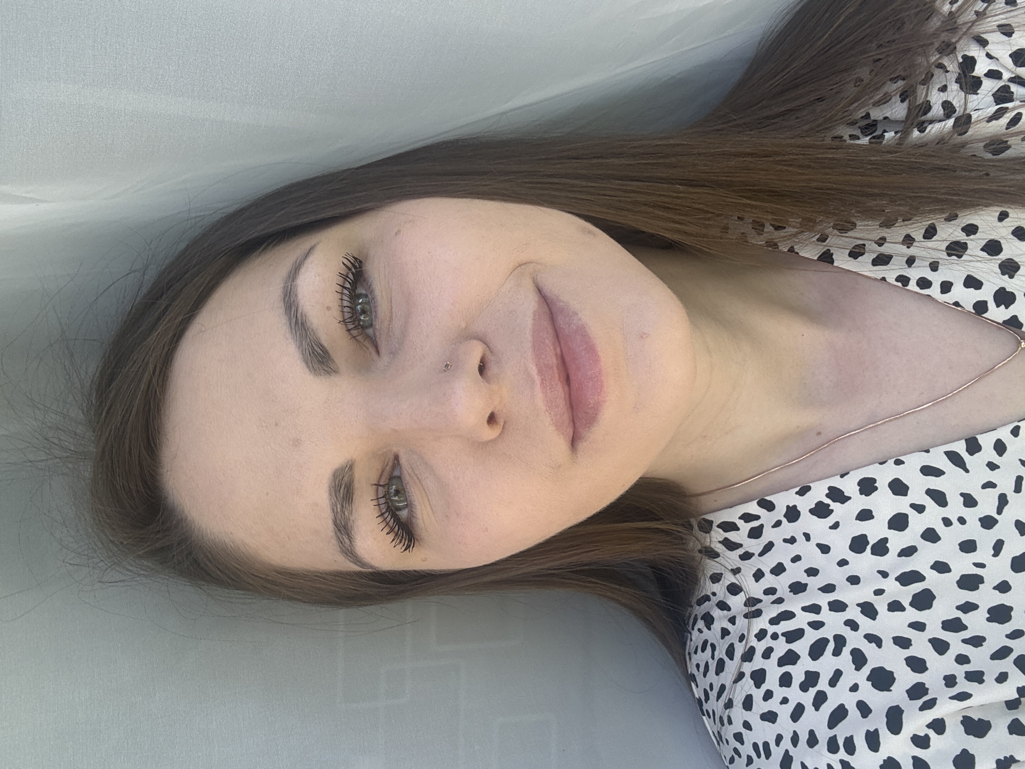 A selfie of a woman with long, straight brown hair is lying down, facing the camera. She has fair skin, light green eyes, and dark eyelashes and eyebrows. She has a nose piercing on the left side of her nose. She is wearing a black and white spotted shirt and a thin gold necklace. The background is a solid light gray surface. The location is unknown.