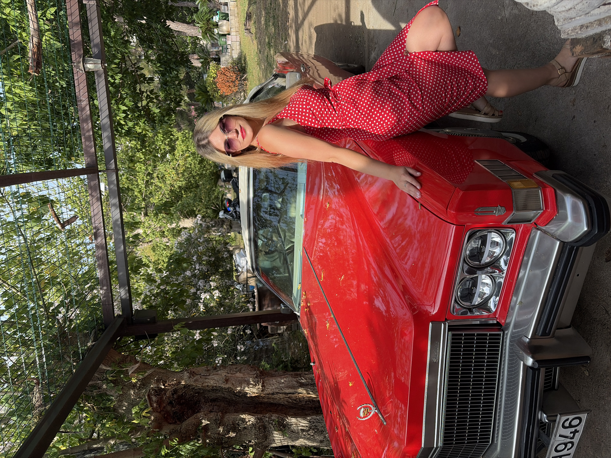 A woman in a red polka-dot dress is leaning on the hood of a red classic car in Budva, Montenegro. She wears sunglasses and sandals. The car has a white convertible top and a license plate. The scene is outdoors with trees and foliage visible in the background and a metal structure to the left.