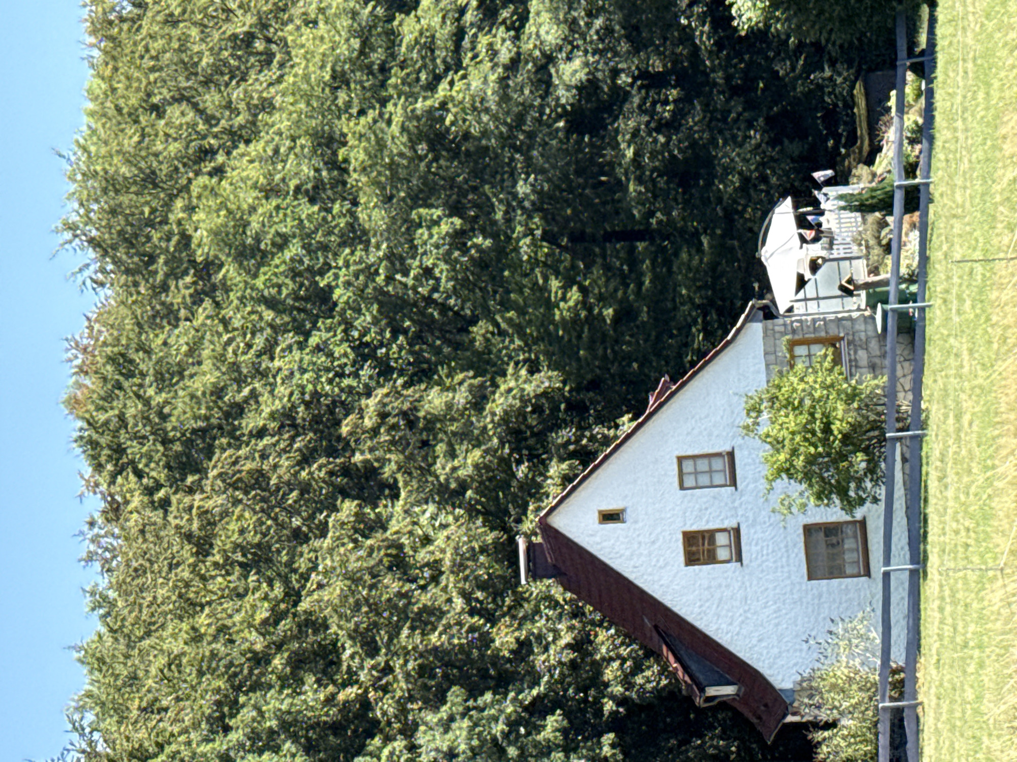 The image shows a white, two-story house with a red roof, situated against a dense backdrop of green trees. The house has several brown-framed windows. In front of the house, a wooden fence runs horizontally across the scene, separating it from a field of green grass. To the right of the house, a white umbrella with decorative flags is set up on a patio area, next to a small green structure. A slender evergreen tree stands near the patio. The sky above is clear and blue. The scene depicts a rural or suburban residential setting. The location is identified as being in the city of Bad Sachsa, Germany.