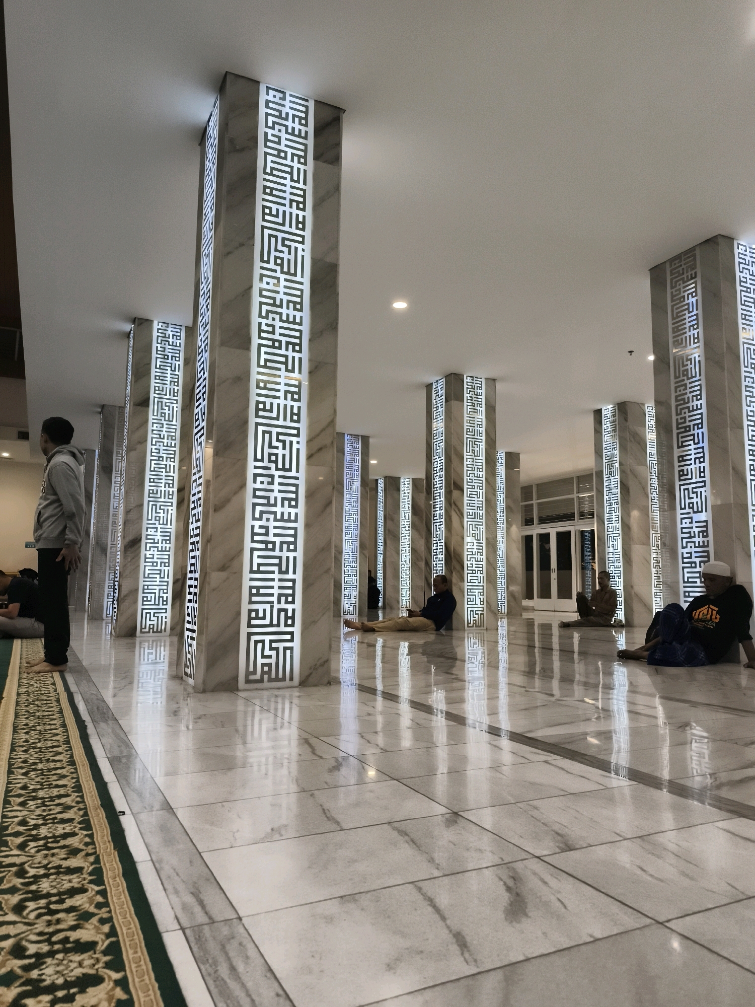 The image depicts the interior of a mosque, likely the Masjid Agung Jawa Tengah in Semarang, Indonesia, given its distinctive architectural features. The space is characterized by a brightly lit, marbled floor that reflects the light from the ceiling and the illuminated pillars. Several tall, rectangular pillars, clad in marble and adorned with intricate Arabic calligraphy in a modern, stylized font, line the hall. These pillars are a central design element, with lights embedded within them, casting a glow that highlights the calligraphy.
In the foreground, on the left side, a man in a grey hoodie and dark pants stands with his back to the camera, looking towards the pillars. To his left, a portion of a green and gold patterned carpet is visible, suggesting an area for prayer. Further back, several individuals are seen seated or reclining on the floor. One person in a blue shirt is sitting with their legs extended, and another in a dark shirt and blue wrap is seated with their legs crossed. In the distance, more figures are visible, some seated, and others appearing to be walking or standing. The background reveals a large doorway with glass panels and doors, leading to an exterior area. The overall atmosphere is serene and contemplative, indicative of a place of worship.