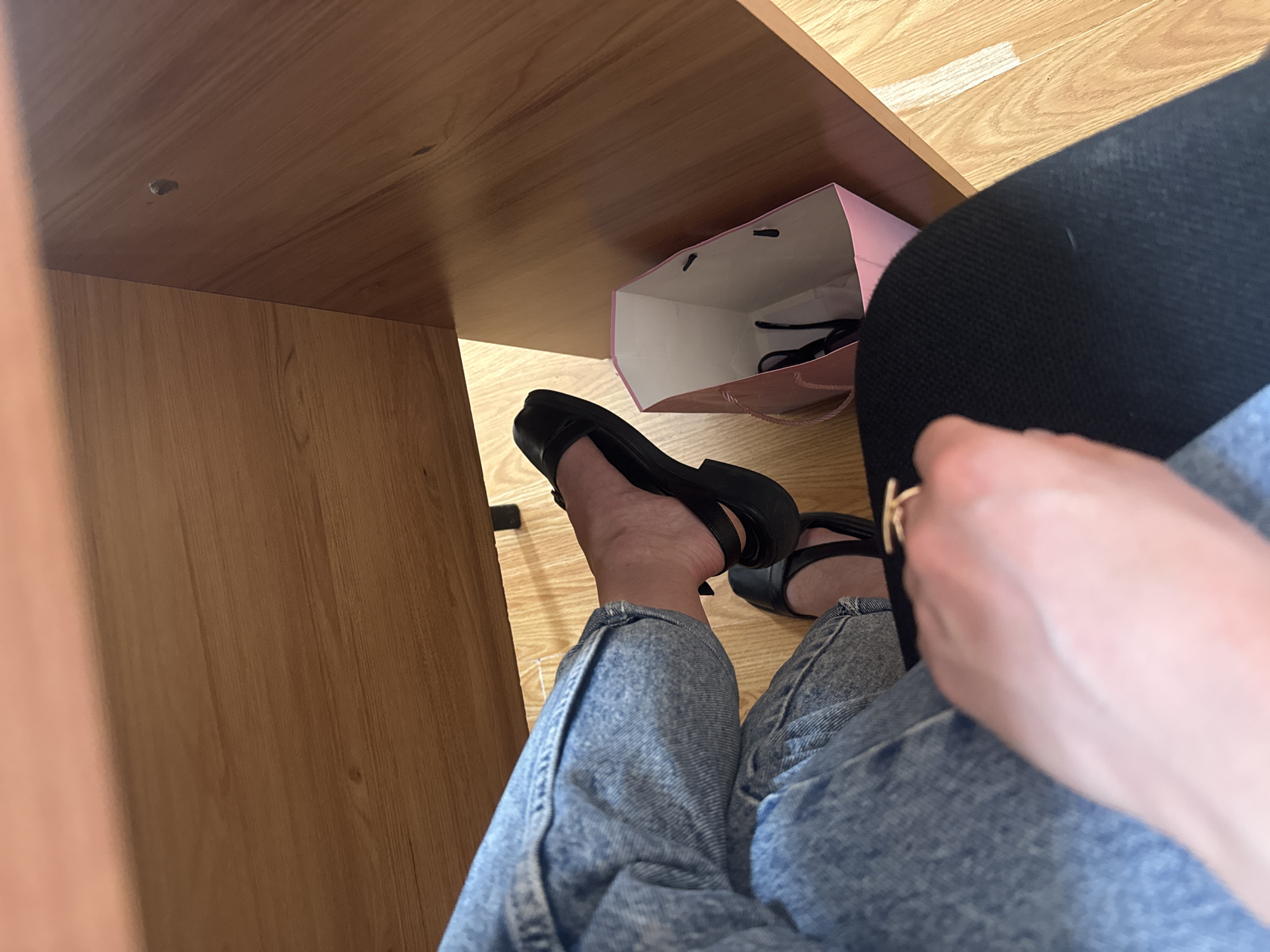 A high-angle shot shows a person sitting with their legs stretched out under a wooden desk. They are wearing blue jeans and black sandals. A pink shopping bag is visible next to their feet, under the desk. The floor is light-colored wood. The location appears to be indoors.