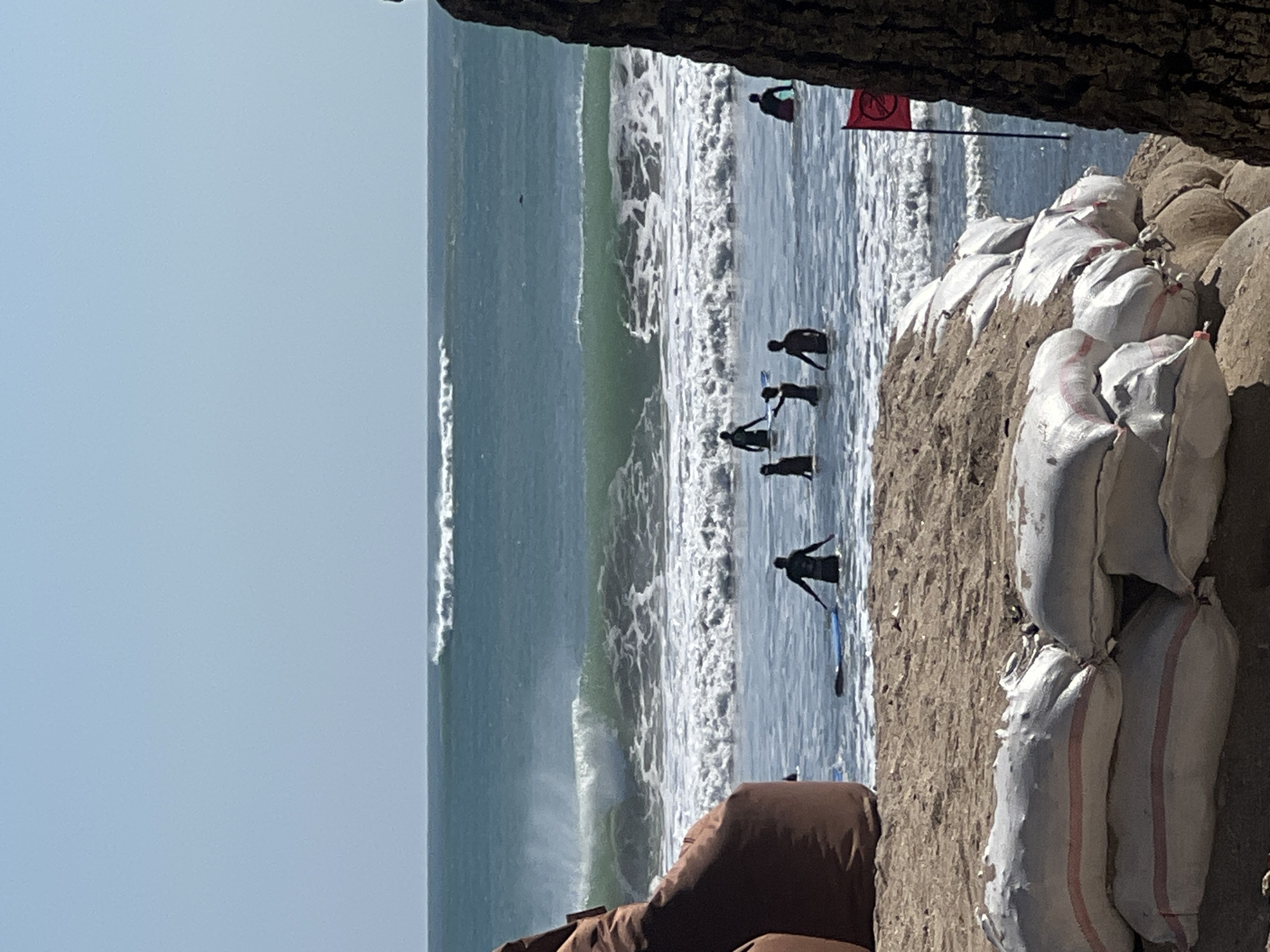 The image depicts a bright, sunny day at a beach in Kuta, Indonesia. The ocean is a vibrant turquoise, with moderate waves breaking and creating white foam. Several surfers are visible in the water, some standing and others with their surfboards. In the foreground, a sandy beach is visible, with large sandbags lined up along the shore, possibly for erosion control. A large brown beanbag chair is partially visible on the left side of the frame, and a tree trunk is on the right. A red flag with a white symbol is planted in the sand near the water's edge, indicating a potential hazard or warning. The sky is clear and pale blue, with no clouds.
