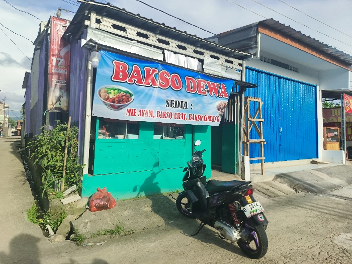 A small, single-story commercial building with a teal facade stands along a narrow street. It is located in Cikalong, Indonesia, at the coordinates -6.751 latitude and 107.445 longitude. A large blue banner with white lettering above the entrance advertises "BAKSO DEWA," and a smaller sign below lists the food items available: "Sedia: Mie Ayam, Bakso Crat, Bakso Cicang." A bowl of noodles is pictured on the banner. A black motorcycle is parked in front of the building. It has a license plate and an unidentified object tied to the rear passenger seat. A small red bag is visible on the ground in front of the building. A blue corrugated metal garage door stands to the right of the storefront, partially obscured by a wooden ladder. The pavement of the narrow street is dirty and cracked. Lush green plants are visible next to the building. The sky is bright blue with a few scattered clouds.