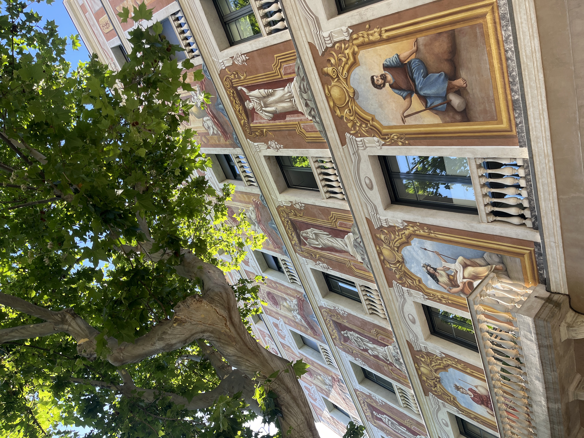 The image shows a multi-story building with ornate murals painted on its facade. The murals depict various figures, some in classical attire, and are framed by decorative gold borders. The building has several windows with black frames and balconies with white balustrades. A large tree with green leaves partially obscures the upper portion of the building. The scene is brightly lit, suggesting it is daytime. The style of the architecture and the murals suggest a location in a European city with a rich artistic heritage. This building is located at Plaça de Catalunya, Barcelona, Spain.