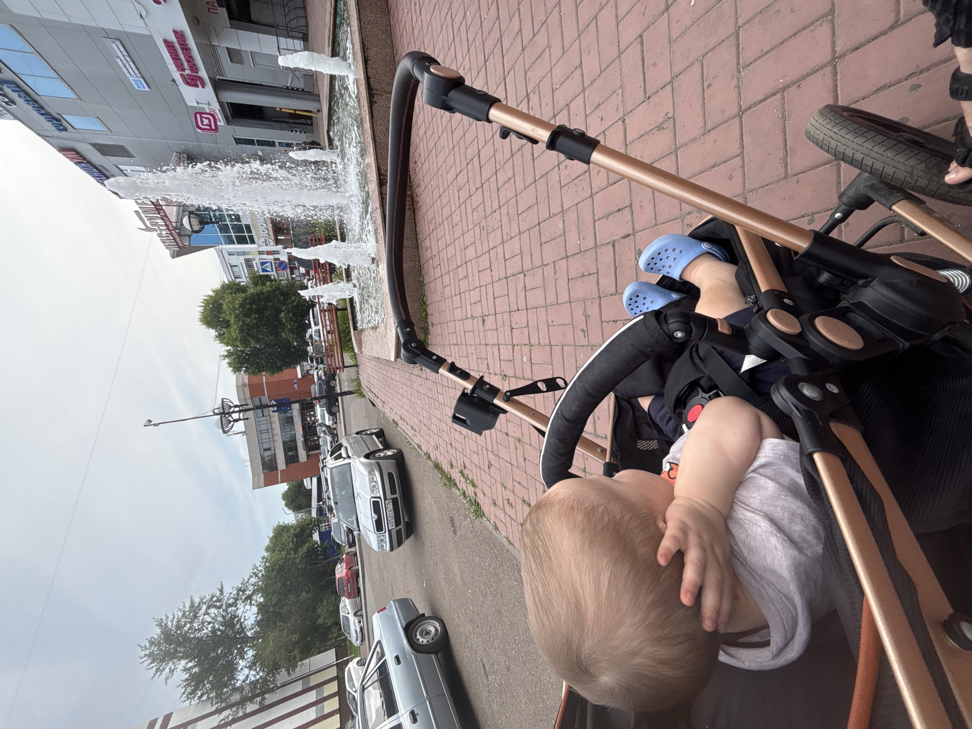 A young child wearing blue Crocs sits in a stroller, resting their head on their arm. The stroller has bronze-colored metal frame components and black fabric. In the background, a street scene unfolds with parked and moving cars, including a silver sedan and a light blue car. A building with a fountain in front of it is visible, and the pavement is made of red bricks. The scene appears to be in Naberezhnye Chelny, Russia.