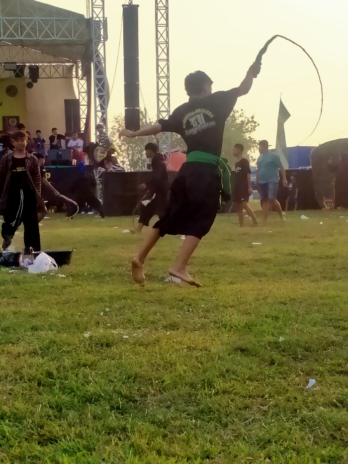 An outdoor scene in Glonggong, Indonesia, features a large grassy field with multiple individuals. In the center, a male performer, viewed from behind, is captured mid-leap with both bare feet off the ground. He wears a dark short-sleeved shirt with white text reading "KESENIAN BARONG DOR" and black pants, cinched with a green sash around his waist. His right arm is extended upward, holding a long, flexible whip that forms a circular arc above him.
To the left in the foreground, another male stands near a black basin containing white, lumpy material and white fabric scraps. In the background, a large metal truss stage structure is visible, equipped with prominent dark speakers. Several individuals, primarily males, are gathered on and around this stage, some appearing to be spectators or additional participants. Other males are distributed across the grassy field, including one in a light blue shirt and shorts on the right side. The ground is predominantly green grass, with scattered white debris. The sky is a light, hazy yellow, indicating soft natural light.