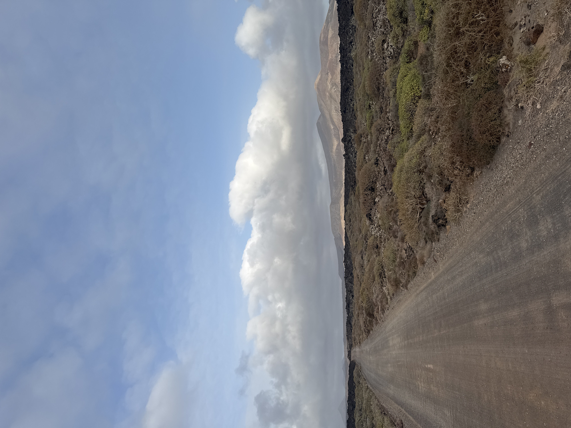 The image depicts a landscape near the city of Tinajo, Spain. A dirt road stretches into the distance. The sky is visible to the left of the road and has white clouds with a blue background. To the right of the road is a strip of land with vegetation that appears dry. Behind the strip of land is a mountain ridge. No people or animals are visible.