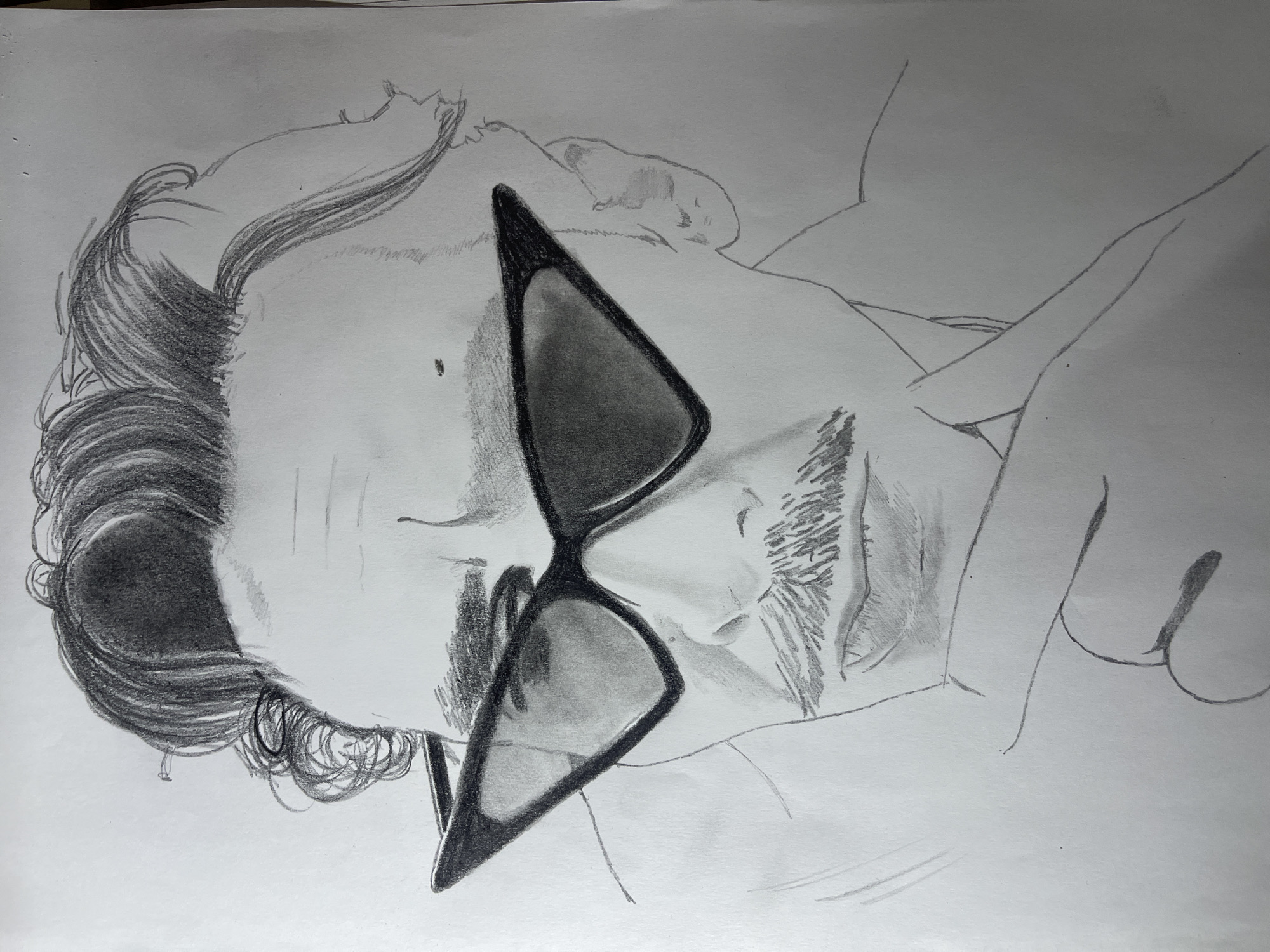 The image is a pencil drawing on white paper of a person lying down. The person has dark, wavy hair, facial hair including a mustache and beard, and is wearing sunglasses. The person's head rests on what appears to be a pillow or soft surface. The upper body is also visible. The background is plain white. There is no indication of the city of Jos, Nigeria, within the drawing.