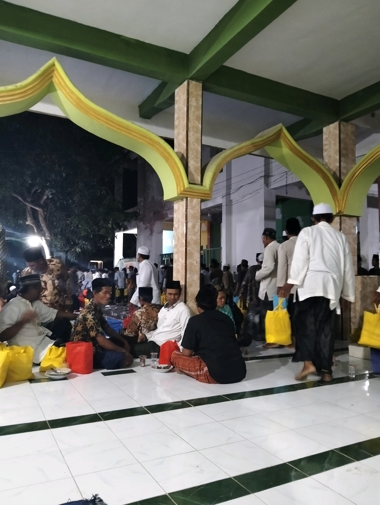 The image depicts an outdoor gathering of people, predominantly men, in Gelap, Indonesia. The setting appears to be a courtyard or open area adjacent to a building with distinctive green and yellow arched structures, possibly part of a mosque or community center. The ground is tiled in white with dark green accents.
Several individuals are seated on the floor, some of whom are wearing traditional Islamic attire, including white caps and long-sleeved shirts. Others are standing and appear to be in conversation or moving through the gathering. Yellow shopping bags are visible in the foreground and middle ground, suggesting that food or goods may have been distributed or are being carried.
The lighting suggests it is evening or night, with some artificial lights illuminating the scene, casting shadows and creating a subdued atmosphere. A large tree with dark foliage is present on the left side of the image. The overall impression is of a community event or gathering taking place in a public space.