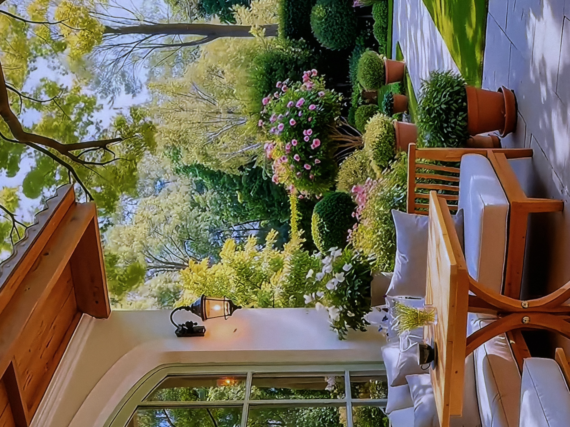 This media file captures a serene outdoor scene, featuring a well-appointed patio adjacent to a vibrant, manicured garden.
In the foreground, a comfortable outdoor seating area is visible, comprising light-colored wooden chairs and a matching table, all furnished with plush white cushions. On the table, a small glass vase holds green foliage, and a dark cup with a saucer is placed nearby. An antique-style black metal wall lamp, with its bulb illuminated, is affixed to a white exterior wall that features an arched window reflecting the surrounding greenery. A wooden roof overhang is partially visible above this wall.
Beyond the patio, a lush garden unfolds, densely planted with a variety of trees and shrubs. Some trees display bright, yellowish-green foliage, while others have darker green leaves. The garden is adorned with numerous well-maintained shrubs, including some shaped into topiaries, and flowering plants showcasing delicate pink and white blooms. Several potted plants, some quite large, are strategically placed along a light-colored paved pathway that meanders through a neatly mown lawn.
The scene depicts a tranquil outdoor space designed for relaxation and enjoyment of nature. No people are present, allowing the focus to remain on the aesthetic appeal of the garden and patio.
The image was captured during a bright, sunny daytime, indicated by the clear shadows and vivid illumination. The overall ambiance is peaceful and inviting. This setting is located in Sofiivska Borshchahivka, Ukraine. There is no visible text in the image.