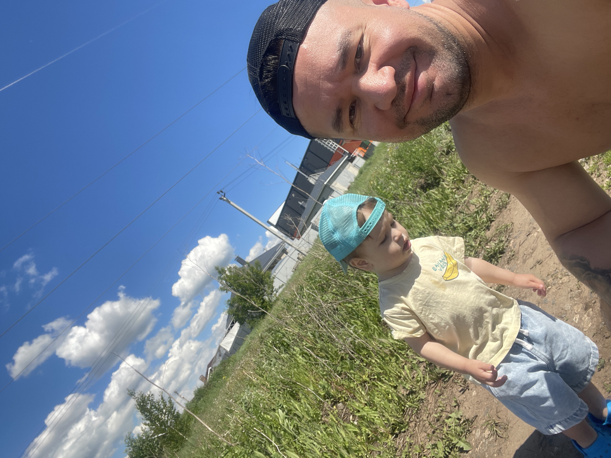The image is a selfie taken at ground level. A fair-skinned adult male wearing a black baseball cap is in the foreground, angled from upper left to lower right. To the right of the center is a fair-skinned child wearing a light blue baseball cap, light yellow t-shirt, and light blue shorts. The child is standing. The background shows a grassy field, a building, power lines, clouds, and a clear blue sky. The scene appears to be outdoors on a sunny day, potentially in a rural or suburban area. Based on the information provided, the location is Эстачи, Russia.