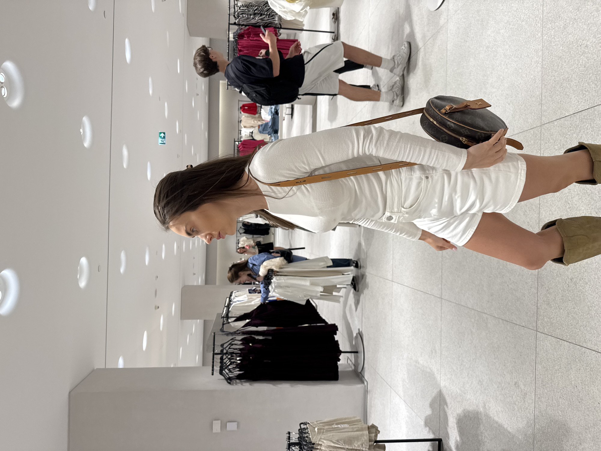 A medium shot of a woman in a white outfit standing in a brightly lit clothing store, her body turned to the right. She wears a white, long-sleeved shirt, white shorts, and brown boots. She carries a brown, rounded bag with a strap across her body. Her dark hair is down.
In the background, a man wearing a black shirt, white shorts, and white sneakers is standing with his back to the viewer. There are clothing racks filled with various garments behind and to the side of the woman. Additional shoppers can be seen in the background as well. The setting is a modern retail space.