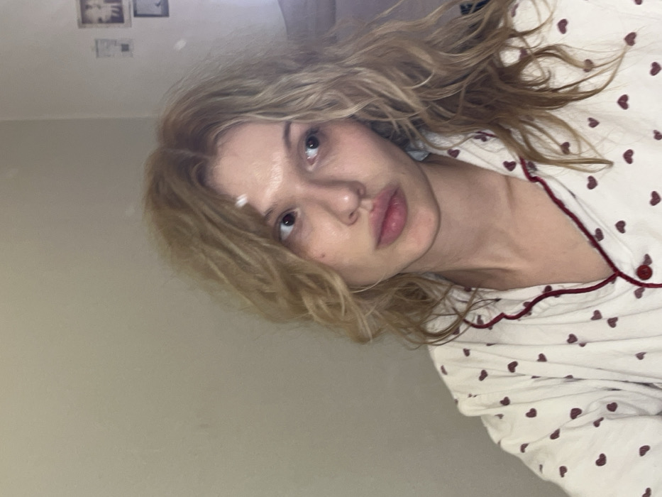 The image displays a young adult female, presenting with light skin and blonde, wavy hair, looking towards the viewer's left. Her hair appears slightly disheveled. She has full lips and discernible dark circles under her eyes; no makeup is apparent. She wears a cream-colored long-sleeved top, possibly pajamas, adorned with small red heart patterns and red piping along the collar and button placket. A single red button is visible near her neck. The background consists of a plain, light-colored wall. In the upper right quadrant, two small, indistinct framed pictures or photographs are mounted vertically on the wall. The scene is indoors, within a domestic setting, located in the city of Chaikovsky, Russia.