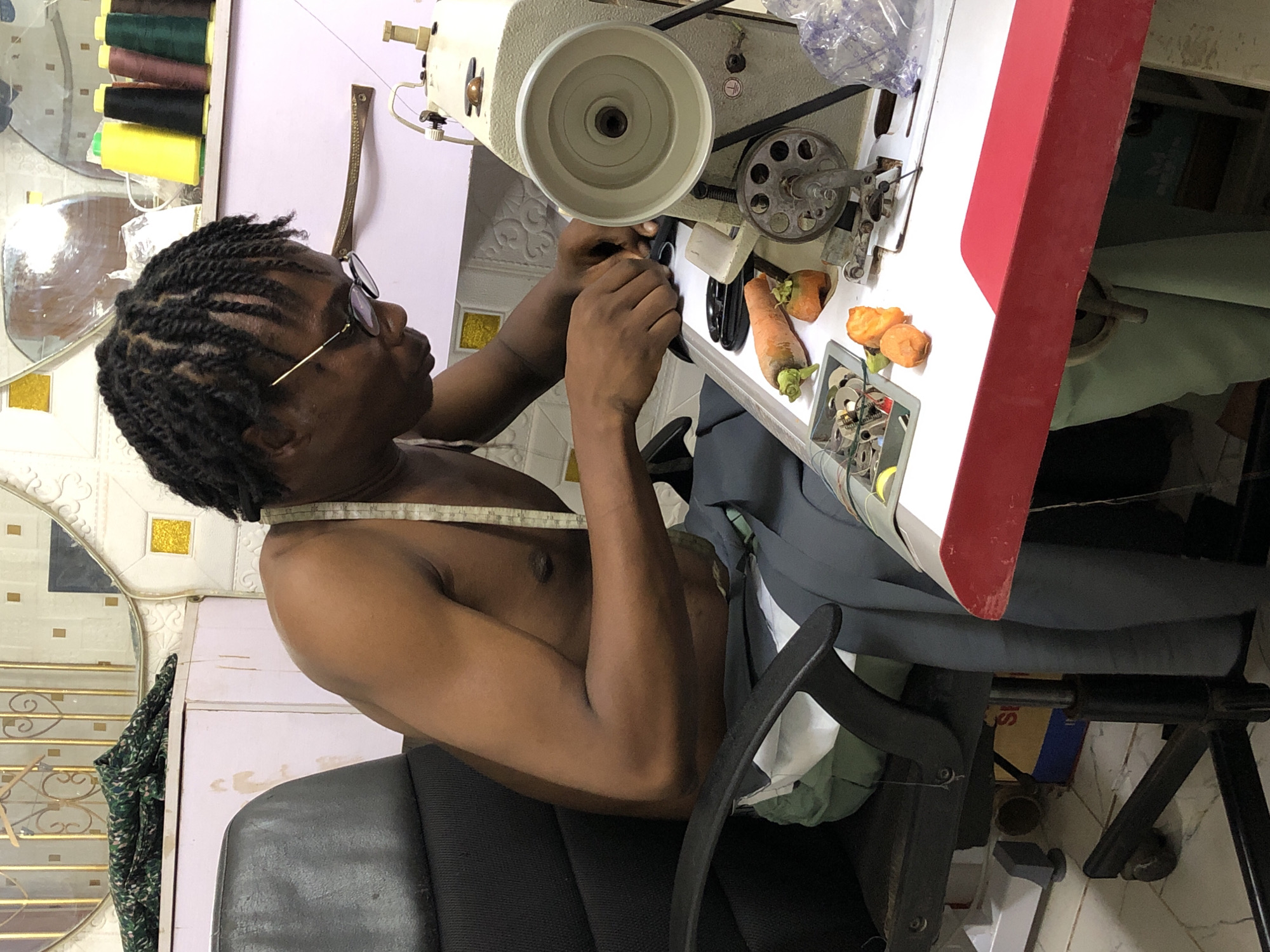 An overhead-view photograph captures an individual operating an industrial sewing machine in what appears to be a tailoring workshop or home studio in Onitsha, Nigeria. The person, with dark skin, wears glasses and has neatly braided hair. They are shirtless, with a white measuring tape draped around their neck, indicating their profession as a tailor or seamster. Their hands are positioned over the machine's needle area, suggesting they are actively sewing or making adjustments.
The workspace is functional, featuring a heavy-duty, cream-colored industrial sewing machine as the central focus. Behind the machine, a rack holds numerous spools of thread in various colors, including brown, dark green, black, yellow, red, and white, signifying a range of tailoring projects. On the white surface of the sewing table, several whole and partially peeled carrots are conspicuously placed, an unusual detail for such a setting. A small grey container nearby holds an assortment of metal objects, likely spare parts, bobbins, or tools for the machine.
The individual is seated on a black, office-style chair. To the left, a mirror reflects portions of a tiled wall with gold accents, hinting at the room's interior design. Fabric, possibly a current project, is draped over the chair and around the workspace. A red and white edge, likely part of the sewing machine table, is visible on the right, and a blue and yellow box can be seen under the table. The lighting suggests daytime. The overall impression is one of dedicated craftsmanship in a modest, practical environment. No discernible text is visible in the image.