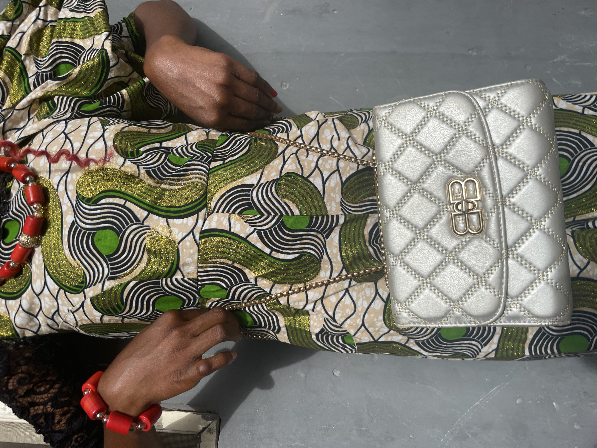 A high-angle shot shows a person, likely lying down, dressed in vibrant African print fabric, with hands and a handbag prominently displayed on a plain grey surface. The fabric features an intricate pattern of green, black, and white wavy lines on a light beige background, with some green sections having a glittery, possibly gold-flecked texture.
One hand, positioned towards the top left, is resting with fingers slightly curled. The other hand, in the lower left, gently grasps the gold chain strap of a handbag. This hand wears a bracelet made of chunky, round red beads interspersed with small silver-colored beads. A similar red beaded necklace is partially visible near the top of the frame. The person's skin tone is dark. Red nail polish is visible on the fingernails of the upper hand.
The handbag, placed on the lower right, is a light silver or metallic grey, quilted, with visible stitching forming a diamond pattern. It has a gold-colored chain strap, parts of which are visible across the fabric. The bag's front flap features a prominent gold-colored metal clasp designed with two stylized, intertwined "B" letters.
The background is a simple, uniform grey surface, possibly a floor, with minor textural variations. The lighting is bright and even, casting subtle shadows. The image captures a detailed view of the attire and accessories. This scene is likely set in Badagry, Nigeria, given the provided context.