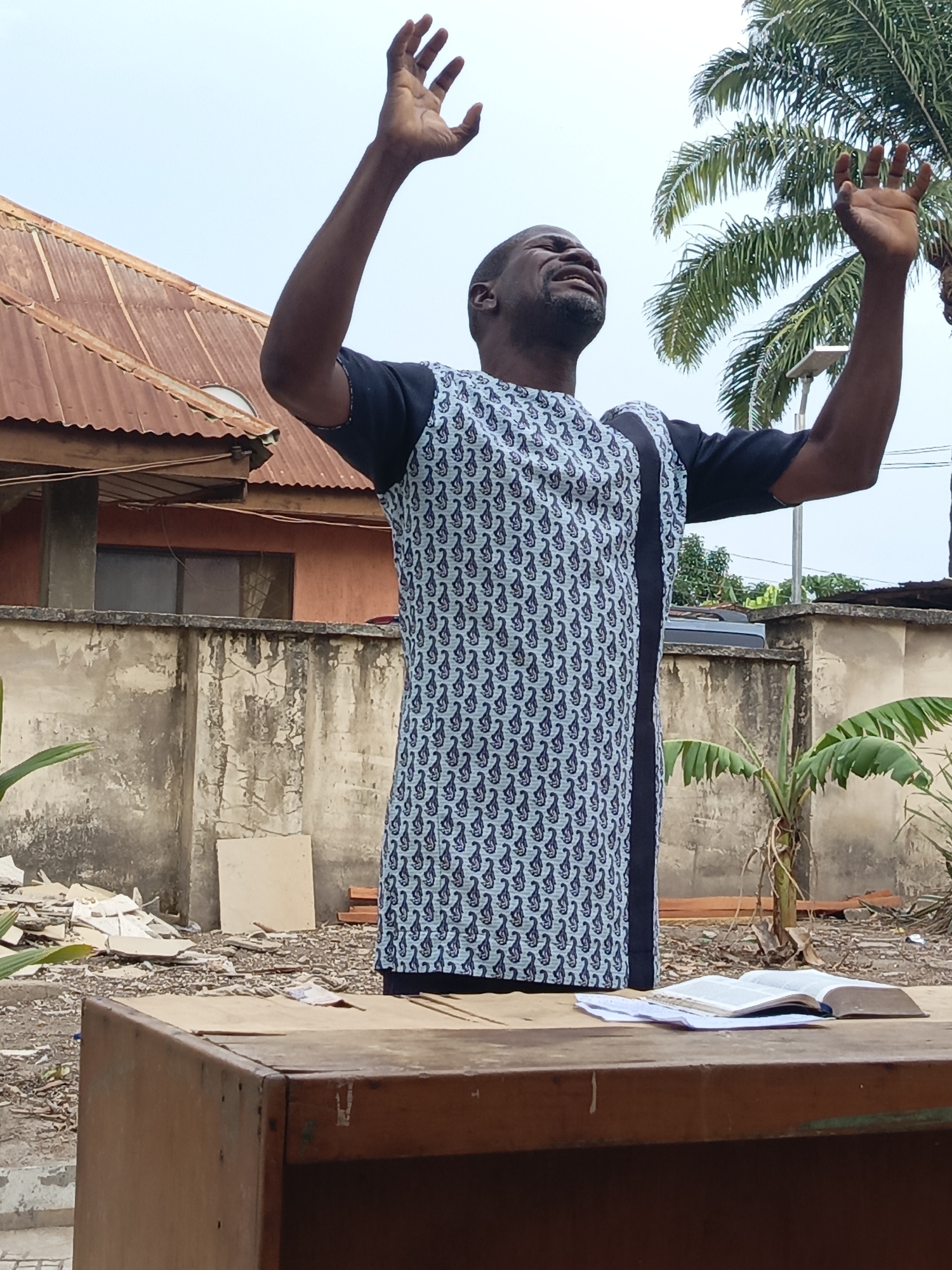 An outdoor scene in Akwanga, Nigeria, captures a dark-skinned man deeply engaged in prayer or worship. He stands behind a rustic wooden desk, with both arms raised high above his head, palms open towards the sky. His head is tilted back, eyes closed, and mouth open, conveying intense emotion and spiritual fervor. He is dressed in a light blue tunic featuring a dark, intricate pattern, with plain black short sleeves.
On the wooden desk in the foreground, a large, open book, identifiable as a Bible, rests alongside some papers. The background reveals an unadorned outdoor setting. To the left, a building with a rusty corrugated iron roof is visible, along with a weathered concrete block wall. To the right, green palm trees and a banana plant add a tropical element. The ground is unpaved, consisting of dirt, gravel, and scattered debris like broken tiles and wood scraps. The sky is overcast, suggesting daytime with diffused lighting. The overall atmosphere is one of humble devotion in a semi-rural or developing residential area.