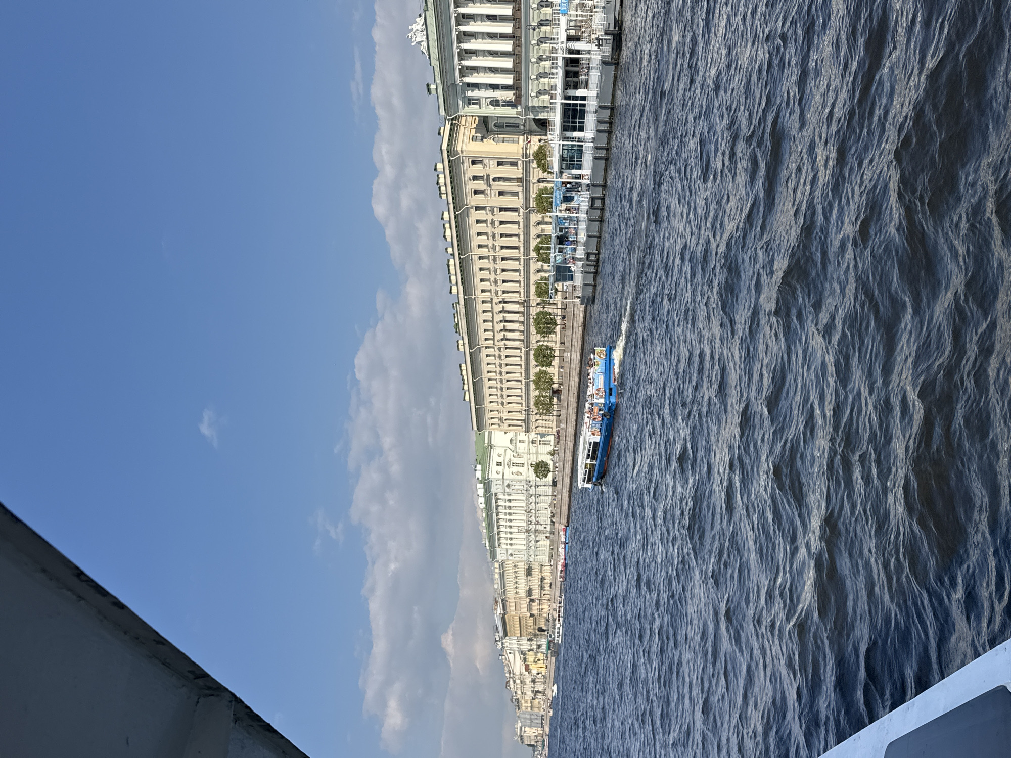 This image depicts a waterway in Saint Petersburg, Russia, with a large, multi-story building lining the embankment on the left. The building is a pale yellow color with numerous windows and architectural details like columns and ornate cornices. Several small trees are planted along the embankment in front of the building.
A white and blue tour boat with people visible on its deck is moving along the water, creating ripples. The water itself is a deep blue, reflecting the sky. The sky is a clear, bright blue with scattered white clouds. The perspective suggests the photo was taken from another boat or a low vantage point near the water.
The scene appears to be from a sunny day, likely during daylight hours, given the bright illumination and blue sky. The overall mood is peaceful and scenic, showcasing a typical view of Saint Petersburg's waterways and its grand architecture. There is no visible text in the image.