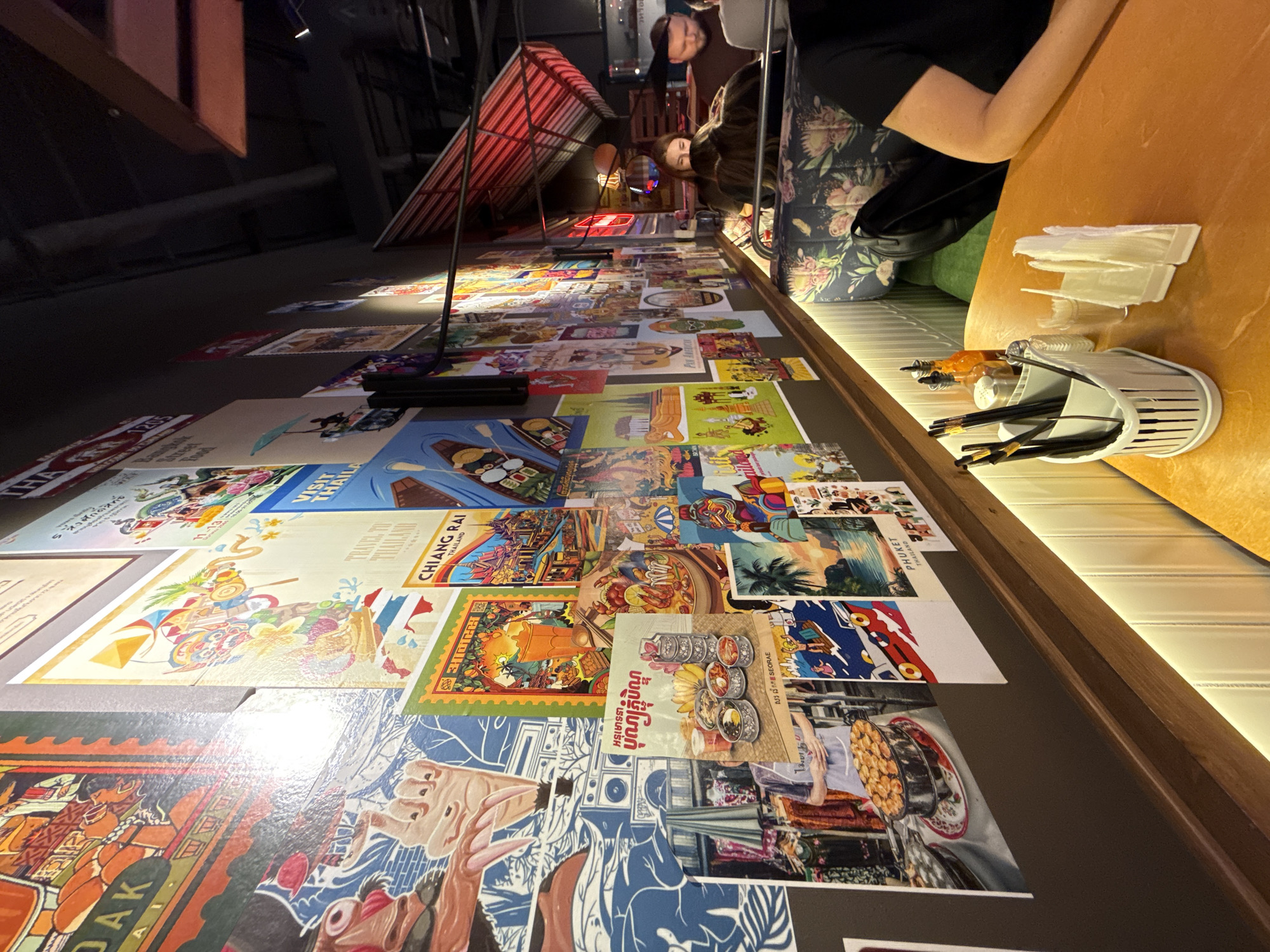 The image shows the interior of a restaurant with walls decorated with numerous posters, many of which appear to be travel posters for Thailand. In the foreground, a table is set with cutlery in a holder, napkins, and condiment bottles. Several people are seated at tables in the background. The lighting is dim, with warm light coming from the table lamps and wall sconces. The overall atmosphere suggests a casual dining environment.