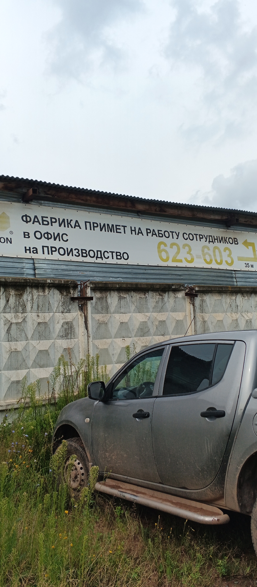 In Pirogovo, Russia, a gray pickup truck is parked amidst tall, green grass and weeds. The truck is positioned on the right side of the frame, with its front partially obscured. Behind the truck, a rough, textured concrete wall serves as a backdrop. Mounted on a corrugated metal structure above the wall is a large, white sign with black and yellow text. The sign reads in Russian: "FABRIKA PRIMET NA RABOTU SOTRUDNIKOV V OFIS na PROIZVODSTVO" which translates to "FACTORY ACCEPTS STAFF FOR WORK IN THE OFFICE for PRODUCTION". A phone number, "623-603", is prominently displayed in yellow on the right side of the sign, with an arrow pointing to the right and the text "35 M" below it. The sky above is overcast with gray clouds.