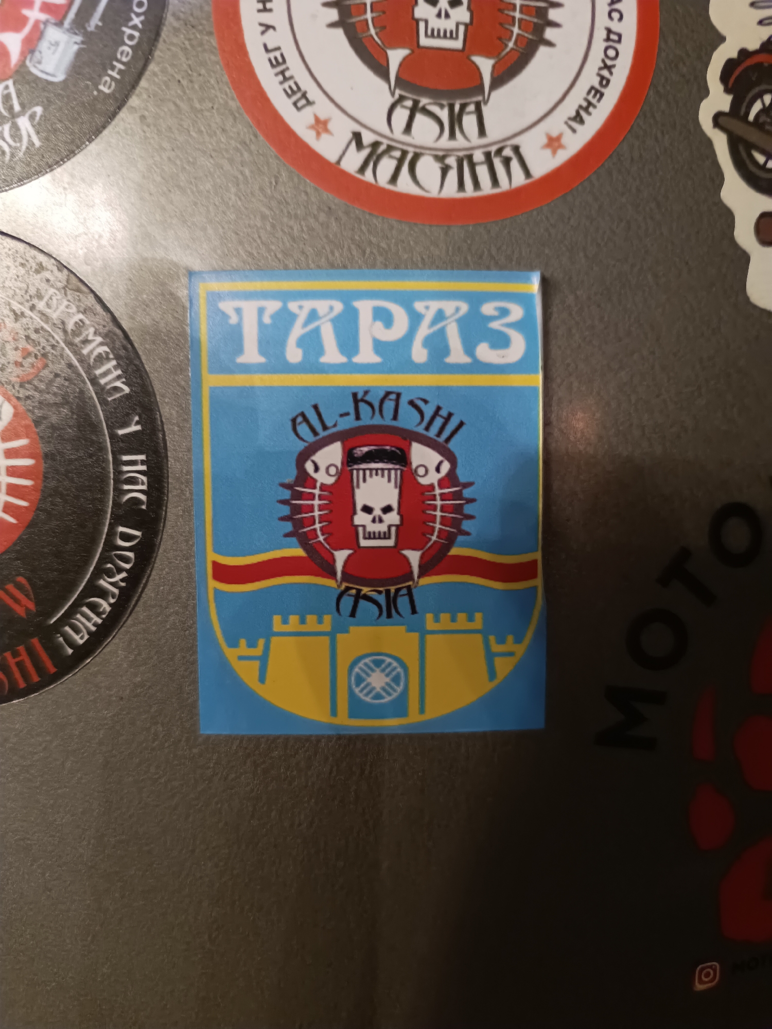 A close-up, eye-level, indoor shot shows a dark, textured surface, likely a refrigerator, with several stickers. A prominent sticker in the center features a blue background with a yellow border and a crest. The crest depicts a stylized red fish with a skull inside, surrounded by smaller fish skeletons and geometric patterns. Above the crest, the word "TARAZ" is written in large, white, stylized letters, and below it, "AL-KASHI" is written in smaller, black letters. The bottom of the sticker shows a yellow castle with two towers and an emblem in the center. To the left and right of the central sticker are circular stickers with text and graphics that are partially visible. The sticker on the left is black with white text that appears to be in Cyrillic. The sticker on the right is red and white with Cyrillic text and an image of a motorcycle wheel. The bottom right of the image shows a dark sticker with the word "MOTO" written in large, white, stylized letters, and a faint Instagram logo is visible at the very bottom. The lighting is dim, casting soft shadows and highlighting the texture of the surface.
