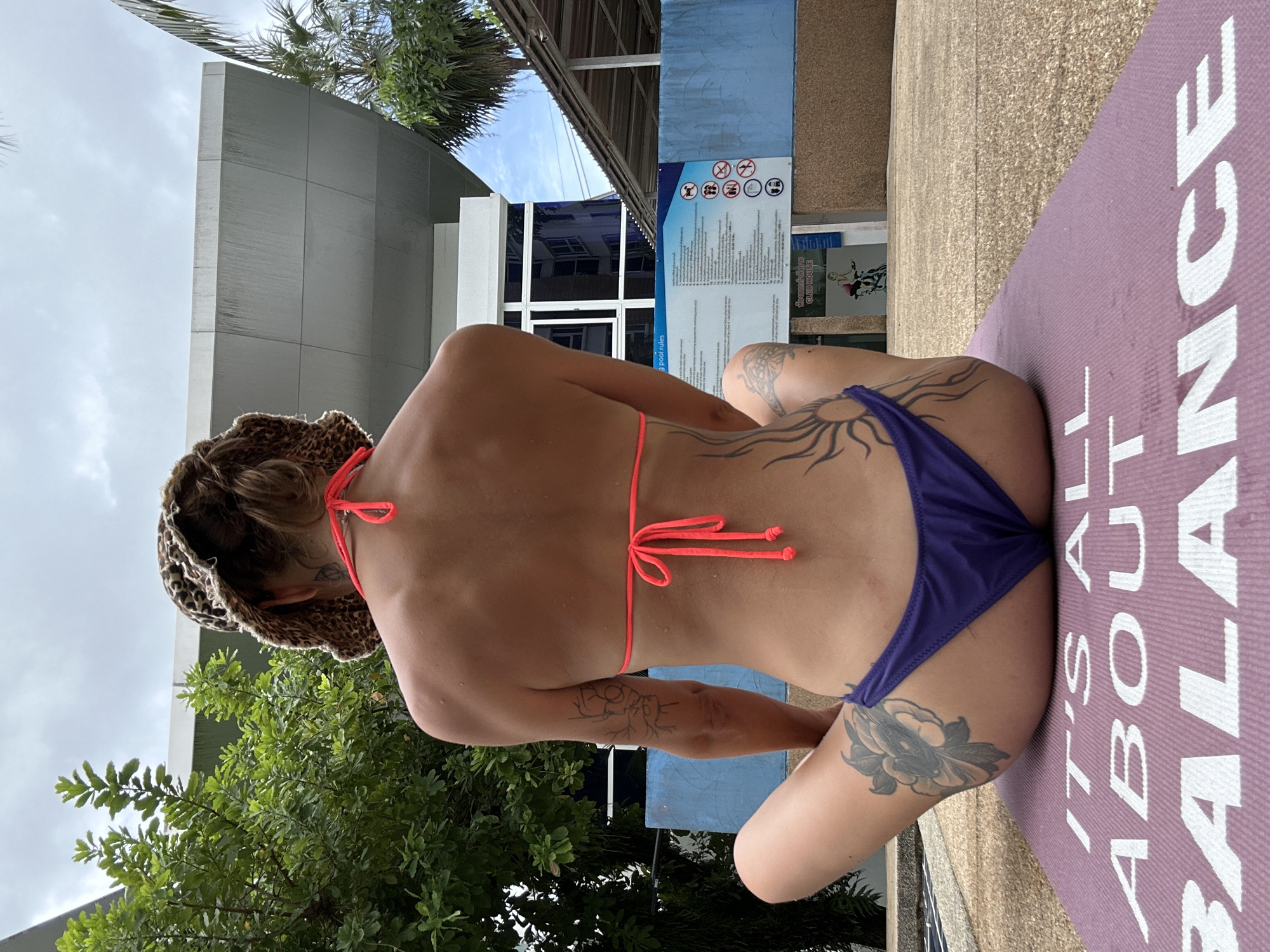 A person is sitting cross-legged on a purple yoga mat in front of a building with blue accents. The person is facing away from the camera and is wearing a leopard print bucket hat, a neon pink bikini top, and purple bikini bottoms. The person has tattoos on their back, arm, and leg. A sign with various symbols and text is visible on the blue wall behind the person. Trees and a palm tree are in the background, and the sky is cloudy. The scene appears to be outdoors, possibly near a pool. The yoga mat has white text that reads "IT'S ALL ABOUT BALANCE." The building in the background may be a club house.