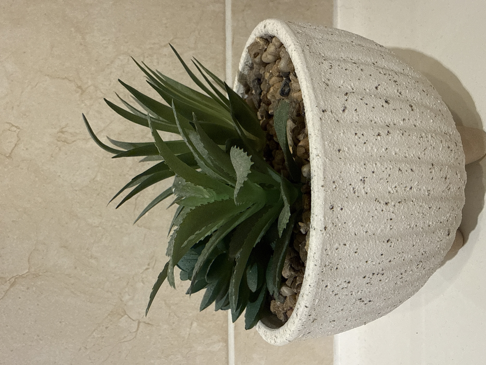 A medium shot depicts a faux succulent plant in a white, textured pot, mounted on a wall. The pot is rounded, speckled with small dark spots, and filled with small brown stones to support the plant. The succulent's leaves are varying shades of green, with some leaves pointing outward and others more closely packed. The background consists of a beige tile wall to the left and a plain white wall to the right. The lighting appears to be artificial. The location is potentially Hazlemere, United Kingdom. No people are visible.