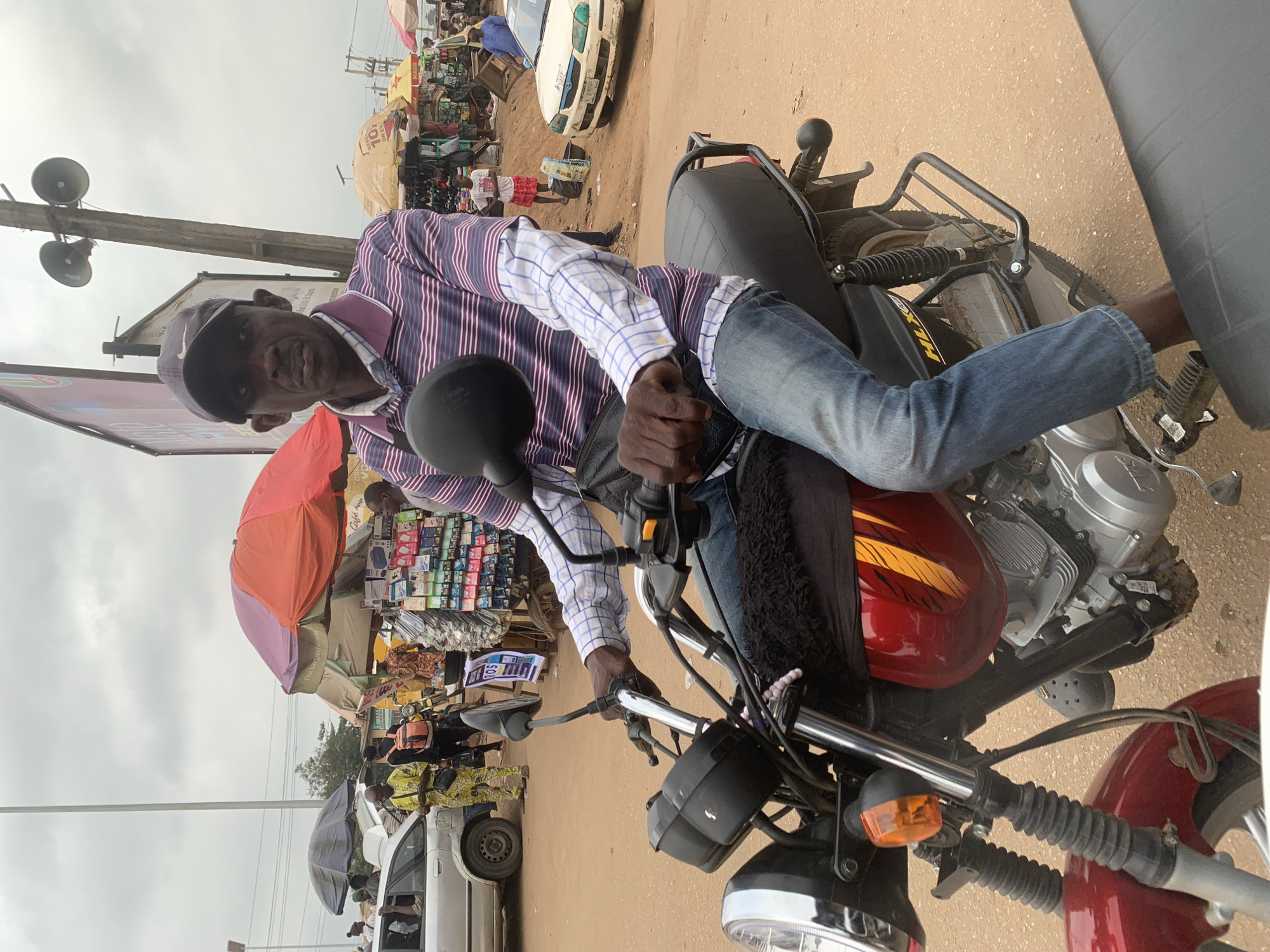 A man wearing a striped polo shirt, blue jeans, and a dark baseball cap sits on a red motorcycle in Shagamu, Nigeria. He is looking forward, holding the handlebars. The motorcycle has chrome accents and a black seat. In the background, a market scene unfolds with various stalls and people. Several umbrellas are set up, providing shade for vendors. A white car is parked on the right side of the street, and other vehicles and individuals are visible in the distance. The ground is covered in reddish-brown dirt, suggesting a dry environment. The sky is overcast.