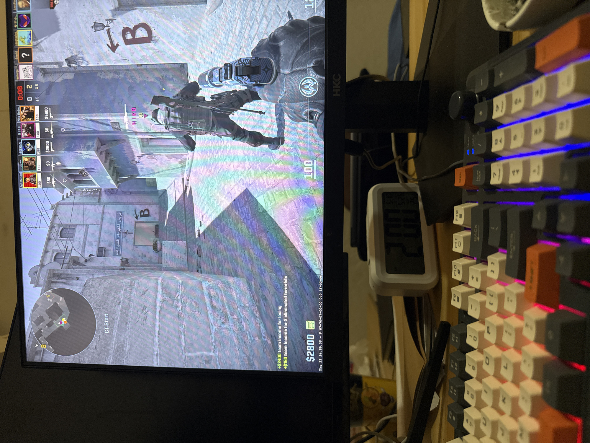 The image shows a computer monitor displaying a first-person perspective video game, Counter-Strike: Global Offensive, being played. The game scene is set in a Middle Eastern-themed environment. The player character, identified as "NIKO" and holding a rifle, is near a building with graffiti "B" and Arabic script. The heads-up display indicates the player's health at 100, the team's money at $2800, and the game time at 0:08. To the right of the monitor is a mechanical keyboard with custom keycaps and backlighting. Other items include a digital thermometer and various cables. The location in Jiulongpo District, China, cannot be determined from the image.