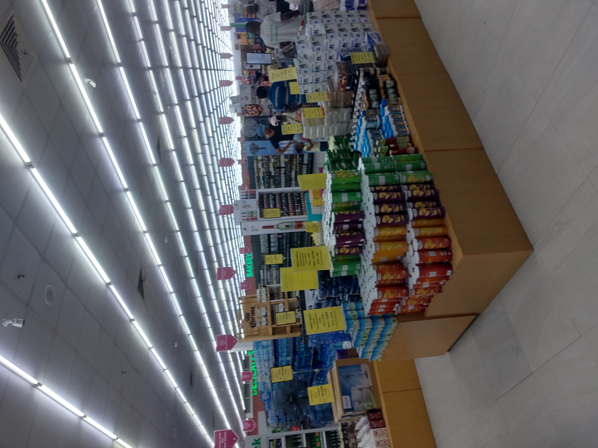 The image displays the interior of a large supermarket, identified as being located in Dar es-Salaam, Tanzania. The expansive retail space is brightly illuminated by numerous parallel rows of white fluorescent tube lights integrated into a grid-style dropped ceiling, which also features a white security camera in the upper left. The floor is covered with light-colored, possibly off-white or light grey, square tiles.
In the foreground and mid-ground, multiple low-profile, light-brown wooden display units are laden with merchandise. Prominently featured are tall stacks of Pringles cans in various colors (red, orange, green, yellow, purple), alongside numerous cans of assorted beverages. Several stacks of white cardboard boxes bearing the "Philips" brand logo are also visible, some likely containing light bulbs or small appliances. Yellow promotional signs, some indicating "GREAT DEALS" and displaying prices in Tanzanian Shillings (TZS) such as "5.100", "2.000", and "59.000", are interspersed among the products.
Approximately seven to nine individuals are present throughout the mid-ground and background, engaged in shopping activities. A man in a dark blue and white striped shirt is partially obscured by a display in the mid-ground. Nearby, a woman wearing a black t-shirt and a light blue cap stands next to a small child in a light-colored top. On the right side, a man in a white t-shirt, possibly a staff member, is seen near a shopping cart. Further into the background, additional individuals are observed browsing aisles.
The background reveals extensive, tall metal shelving units stocked with a wide array of products, including large quantities of bottled water and a dedicated section for alcohol, featuring numerous bottles of wine and spirits. Green-lettered signs indicating "DELICATE" (likely Delicatessen) and "BAKERY" sections are visible. A distinct wooden display unit branded "Corona" is centrally positioned in the background. Red banner-like signs are suspended from the ceiling at regular intervals, suggesting store categories or ongoing promotions.
