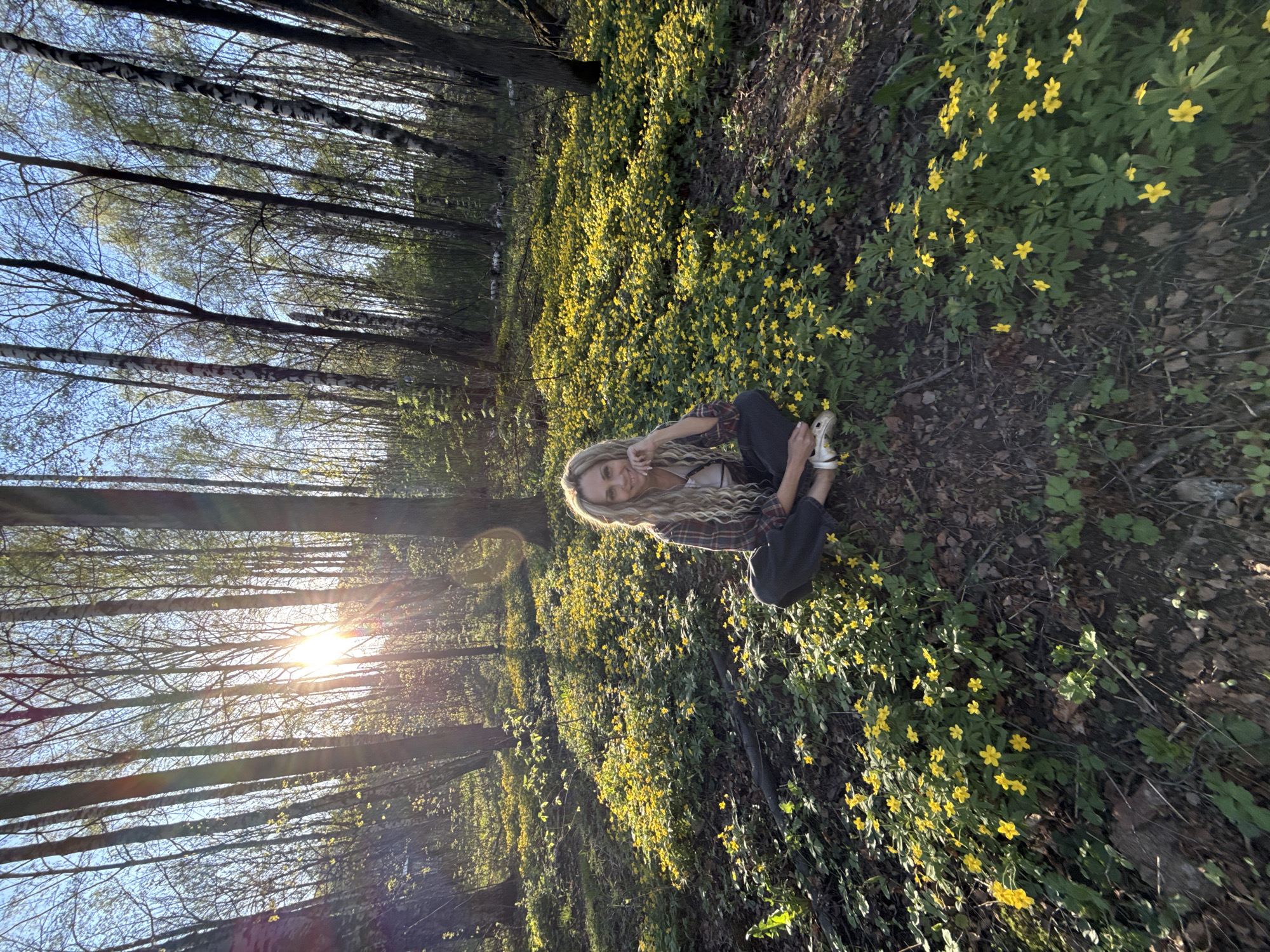 The image captures a woman seated in a forest clearing, which is covered with small yellow flowers. The woman has blonde hair, a light-colored top beneath a flannel shirt, and dark trousers. The sun is shining through the trees in the background, creating a bright light and shadows across the forest floor. The location is Rusanovo, Russia.