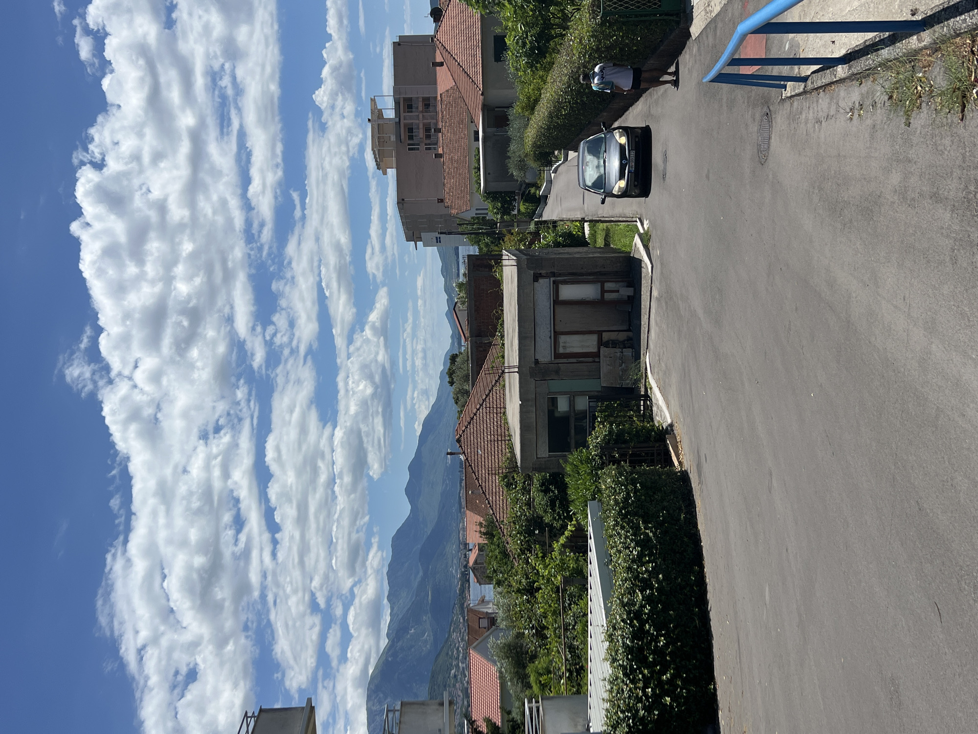 The image depicts a street scene in Bijela, Montenegro, under a partly cloudy blue sky. In the foreground, a paved road curves gently to the right, bordered by manicured green hedges and a blue metal railing on the right side. A man wearing a white t-shirt and dark shorts walks away from the camera on the right side of the road. A dark-colored sedan is driving up the street towards the camera. In the middle ground, several residential buildings with red-tiled roofs are visible, interspersed with lush green trees. A multi-story building with a white top structure stands prominently on the right. The background features a body of water, likely the Bay of Kotor, and a range of distant, mist-shrouded mountains. The scene appears to be a quiet, residential area on a bright, sunny day.