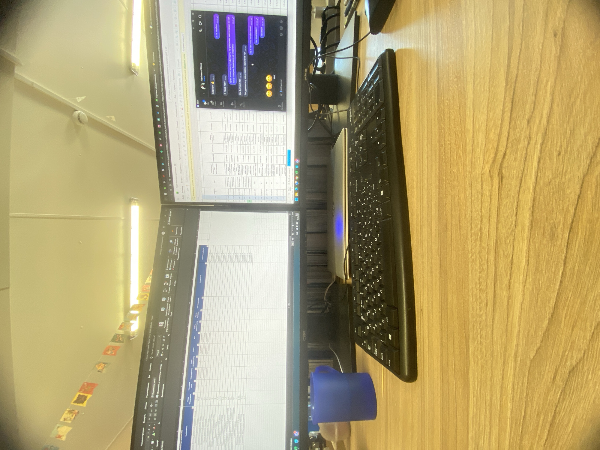 The image shows a desk with two computer monitors, a keyboard, and a blue object (possibly a cup or small container). The monitors display spreadsheets and a messaging application with text in Russian. The desk has a wood grain surface. The image is taken from a slightly elevated angle.
There is no information in the image to indicate the location is Luzhitsy, Russia.