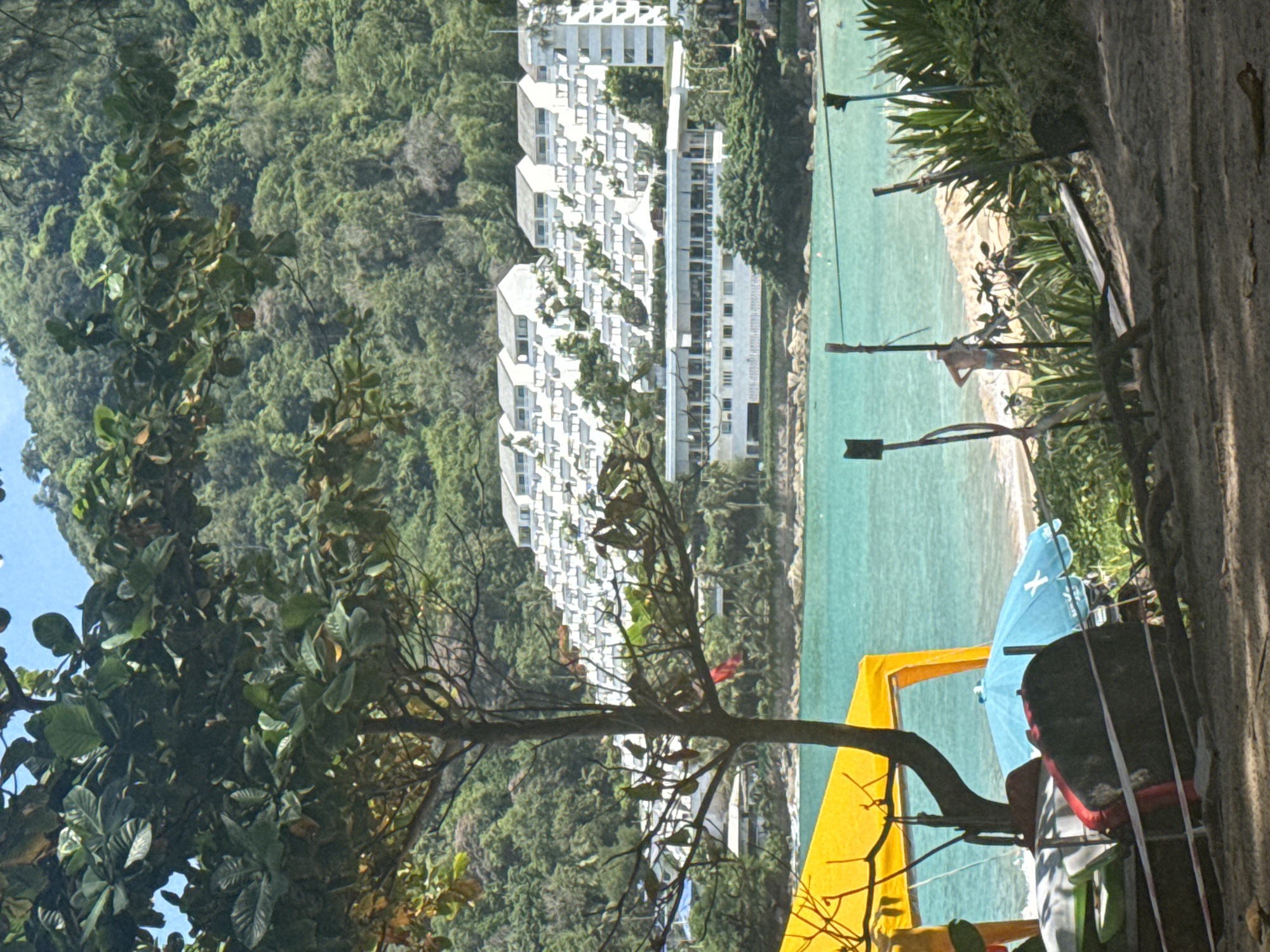 The image depicts a tropical beach scene with a resort complex built on a hillside in the background. In the foreground, a tree with sparse leaves stands to the left, partially obscuring a yellow canopy. Below the canopy, several objects are piled, including what appear to be kayaks or surfboards, a blue umbrella, and a dark, round object. The turquoise water of the sea is visible beyond the beach, and a sandy shore is dotted with vegetation. In the middle ground, a person in blue swim trunks stands on the beach, facing the water. The resort complex consists of multiple white buildings with tiered architecture, integrated into the lush green hillside covered in dense vegetation. The sky is clear and blue.