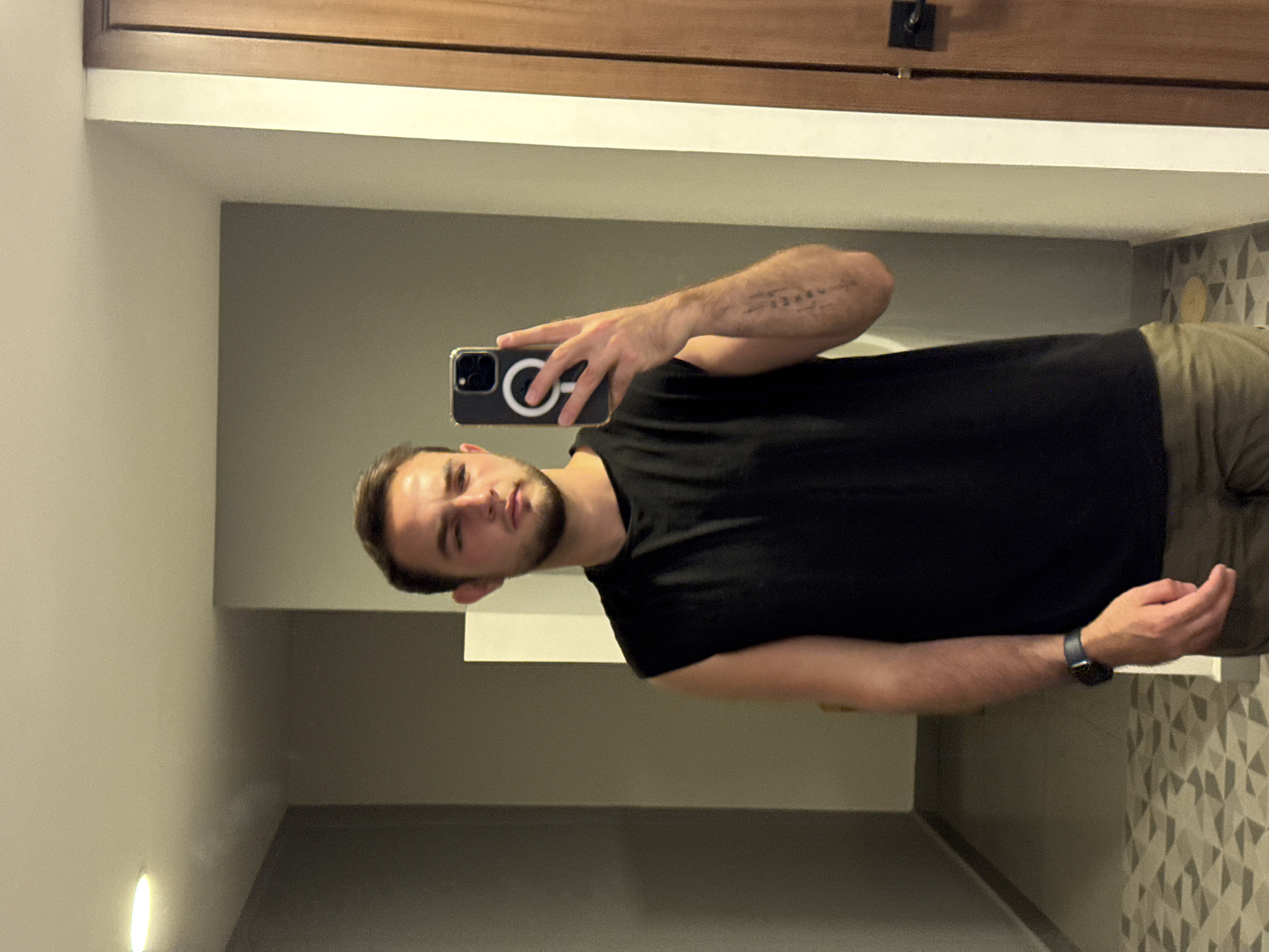 A man is taking a selfie in what appears to be a bathroom. He is wearing a black t-shirt and khaki pants. He is holding a phone with a black case with a prominent white circle design. He is also wearing a dark colored watch on his left wrist. The bathroom has a wood-grain cabinet and a patterned wallpaper. The location is not discernible as San Bernardino Tlaxcalancingo, Mexico.