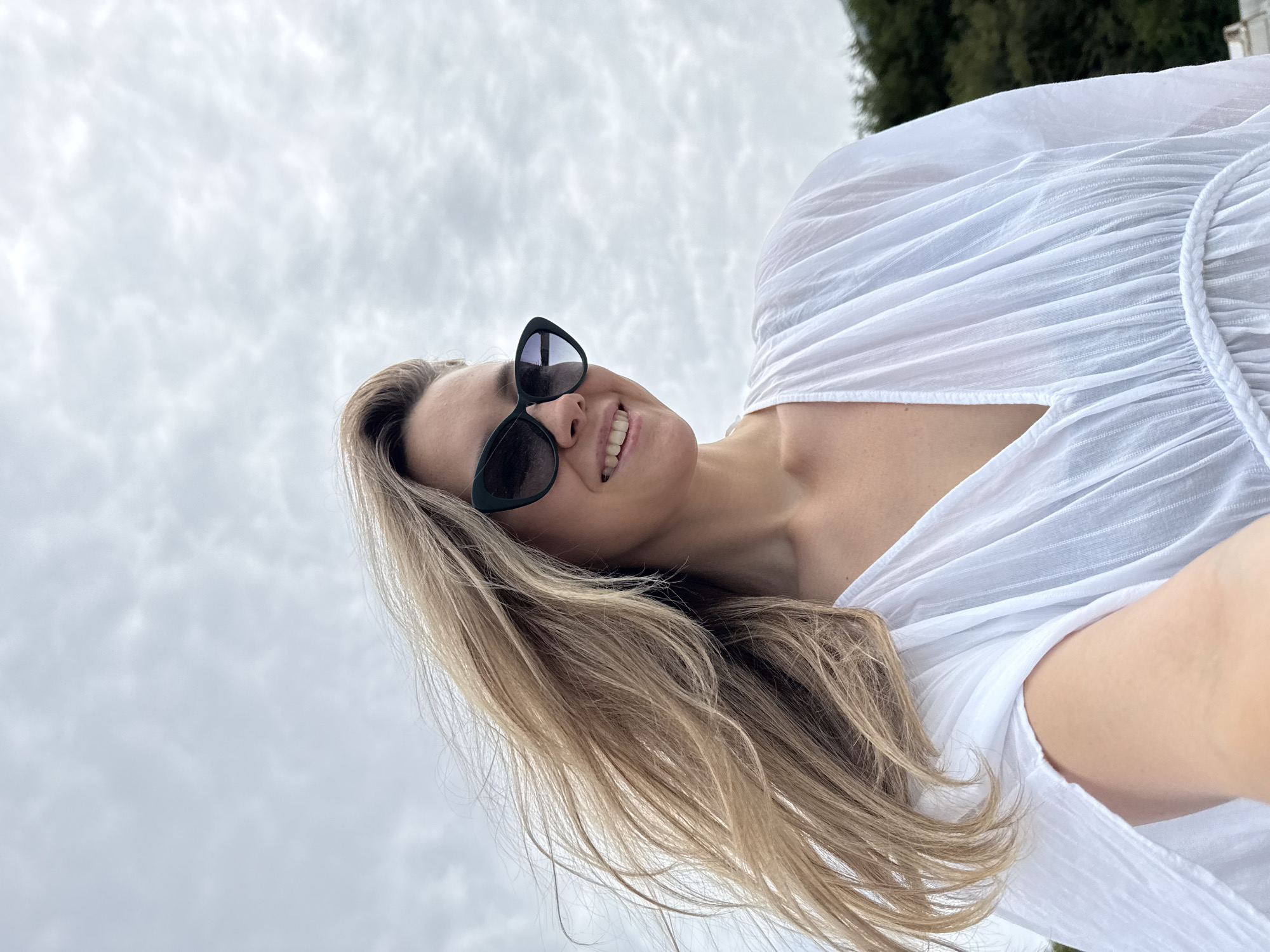 A medium-shot, slightly low-angle, outdoor selfie of a fair-skinned woman with long, blonde hair is captured against a cloudy sky. The woman is wearing oversized, dark cat-eye sunglasses and a white, flowing dress with a V-neck. Her hair is windblown and frames her face as she smiles. The background shows a hint of green trees and what appears to be a cloudy sky typical of Nebug, Russia. The lighting suggests an overcast day.