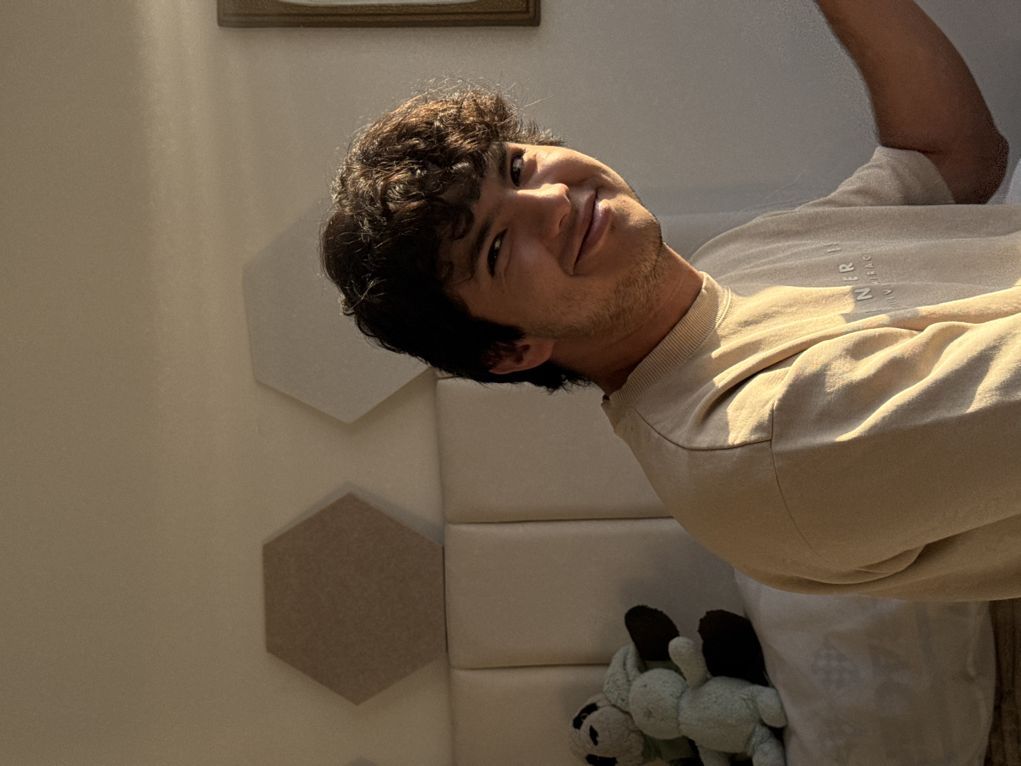 A person with dark curly hair and light brown skin is smiling at the camera. The person is wearing a beige sweatshirt. The background is a white wall with geometric shapes and a framed picture. There are two stuffed animals in the bottom right corner of the image. The location is likely indoors. There is no indication of the city of San Francisco Ocotlán (Ocotlán), Mexico.