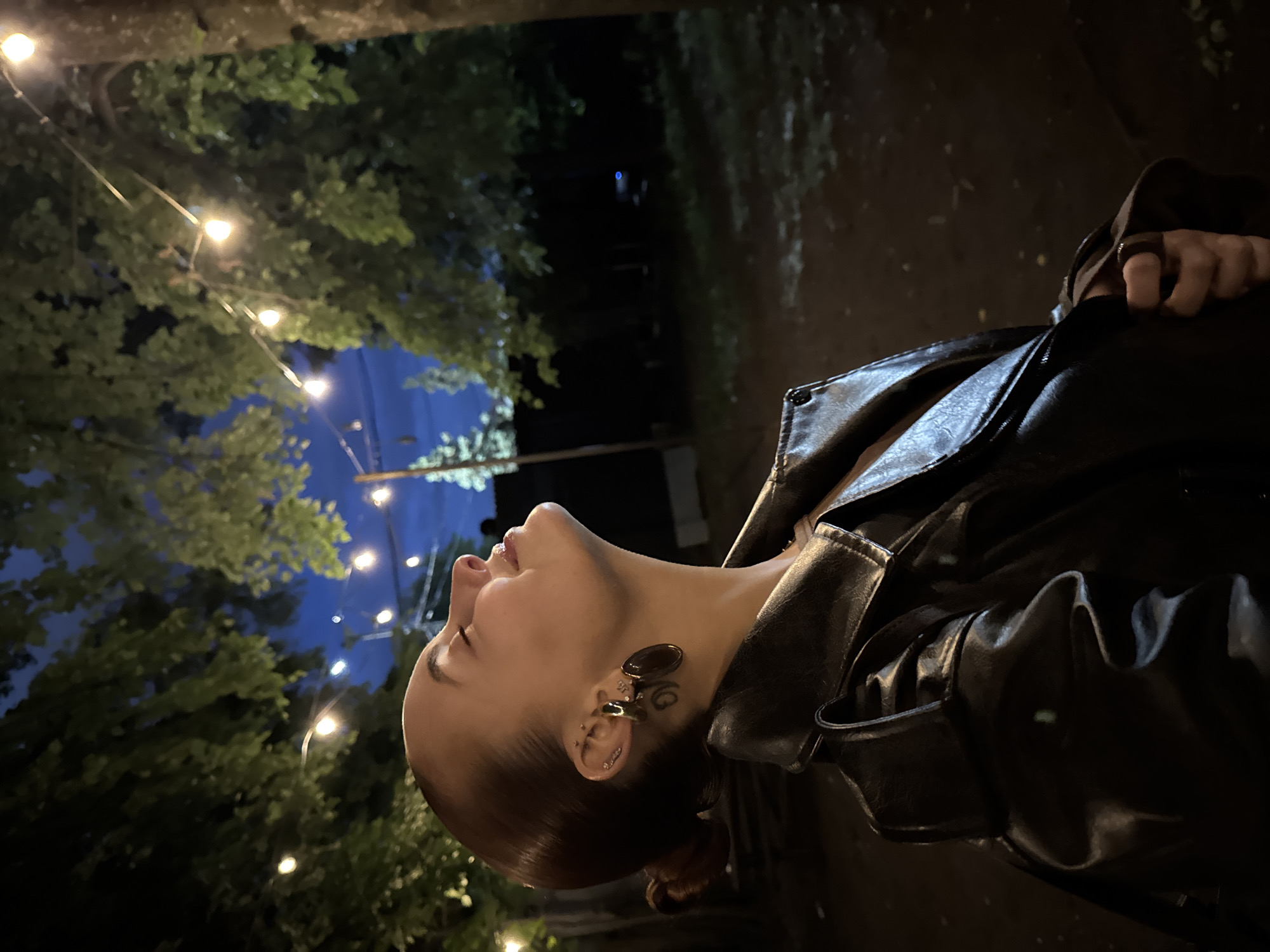 A side profile photograph, taken at night, captures a person (likely female) looking upwards with her head tilted back and eyes gently closed or lowered, suggesting contemplation or peace. She is positioned on the right side of the frame and has dark hair pulled back, revealing a prominent, large, dark, oval-shaped earring in her right ear, along with several smaller piercings. An intricate dark tattoo or design is visible just behind her earlobe. She is wearing a shiny black leather jacket that occupies a significant portion of the lower and right side of the image, with her hand, adorned with a ring, visible near the jacket's collar.
The background depicts an outdoor setting, dominated by dark green foliage of trees against a deep blue night sky, fading into darker tones. Strands of warm-toned string lights are strung between the trees, casting a soft glow and creating an ambient atmosphere. The ground or path in the foreground is dark and possibly reflective. The overall mood is serene and atmospheric, characteristic of an evening spent outdoors under decorative lighting in Izhevsk, Russia. There is no visible text in the image.