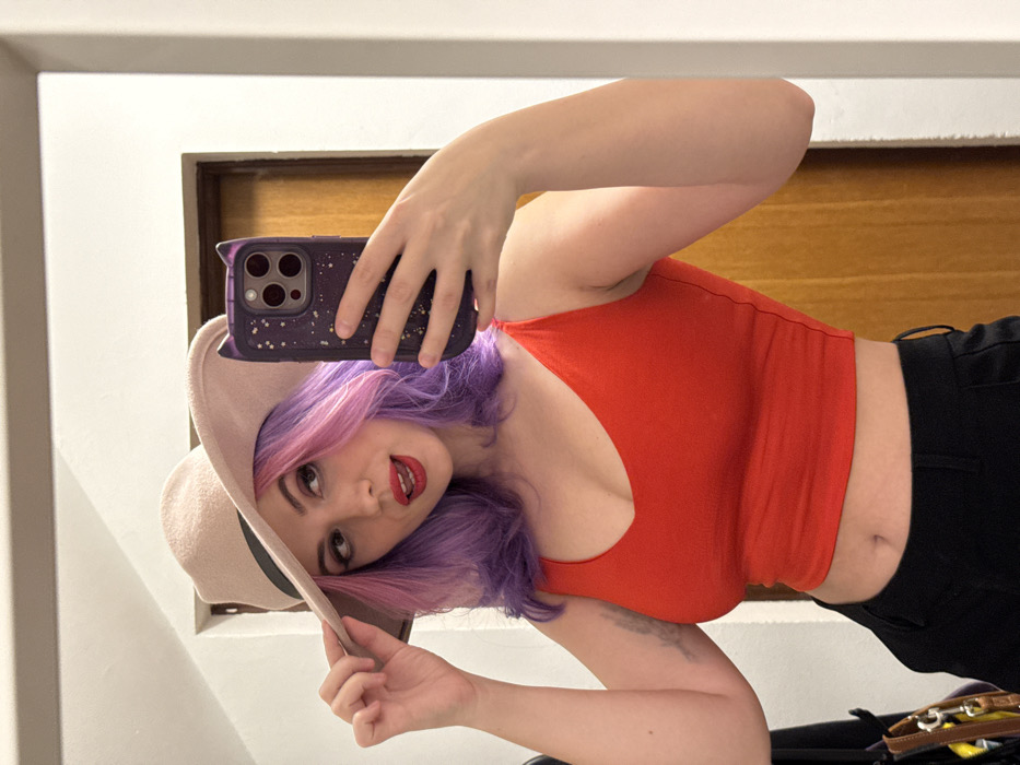 A selfie featuring a woman with purple and pink hair, wearing a beige hat and a red sleeveless top. She holds a dark-colored phone with a speckled case in front of her face, capturing the image. She is posed at an angle, looking at the camera with a slight smile and red lipstick. The background includes a light-colored wall and a wooden panel. The bottom portion of her outfit is black pants.