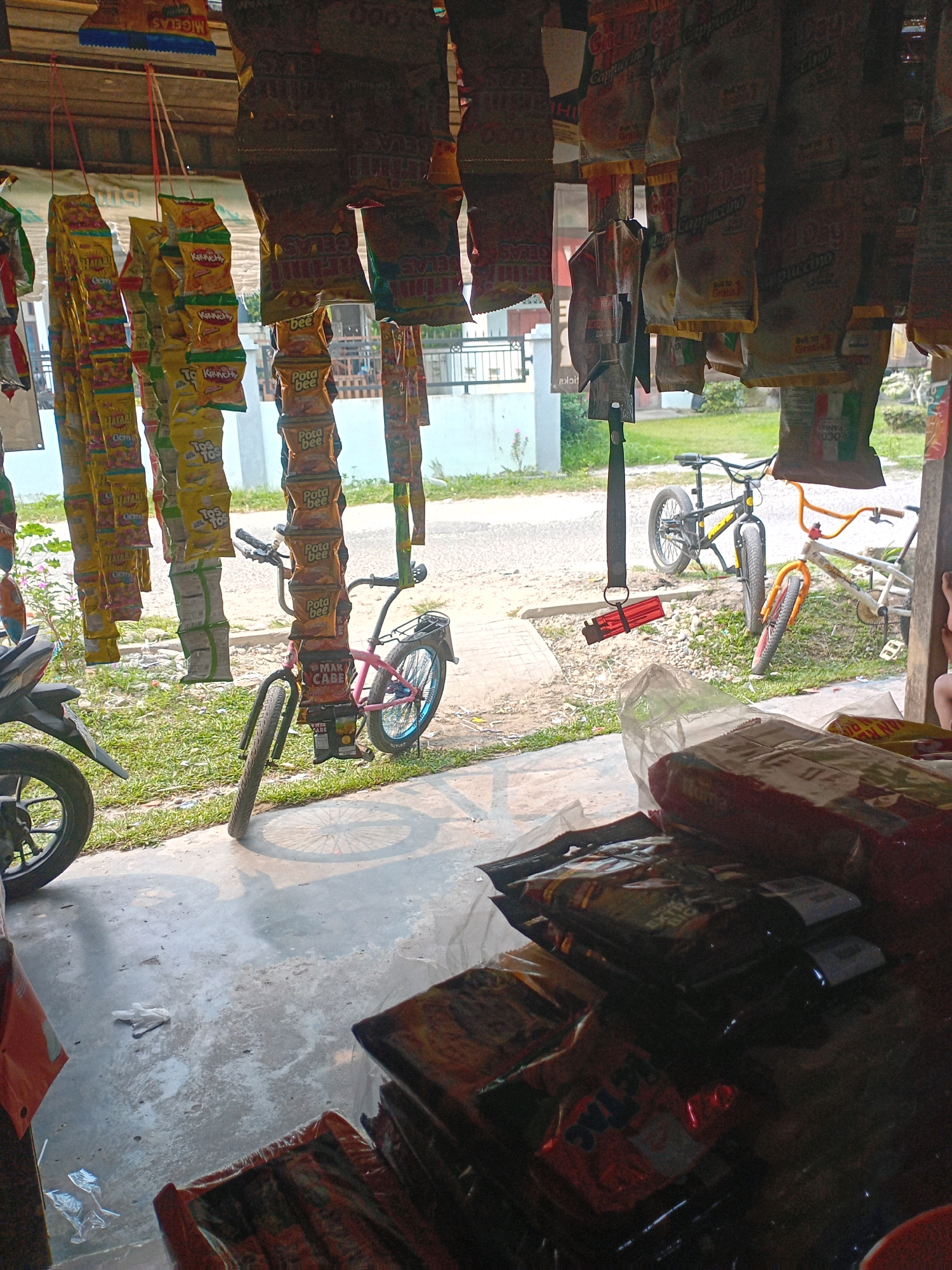 The image depicts a street-side stall in Pekanbaru, Indonesia. The stall is laden with various packaged goods, predominantly snacks and possibly beverages, hanging from the ceiling. In the foreground, stacks of bagged items are visible on a counter or shelf. Two bicycles are present, one of which is leaning against a display of snacks. A motorcycle is partially visible on the left side of the image. The environment suggests an outdoor market or roadside vendor setup, with a concrete floor and an open-air structure providing shade. The lighting indicates it is daytime. There are no people clearly visible in the scene.