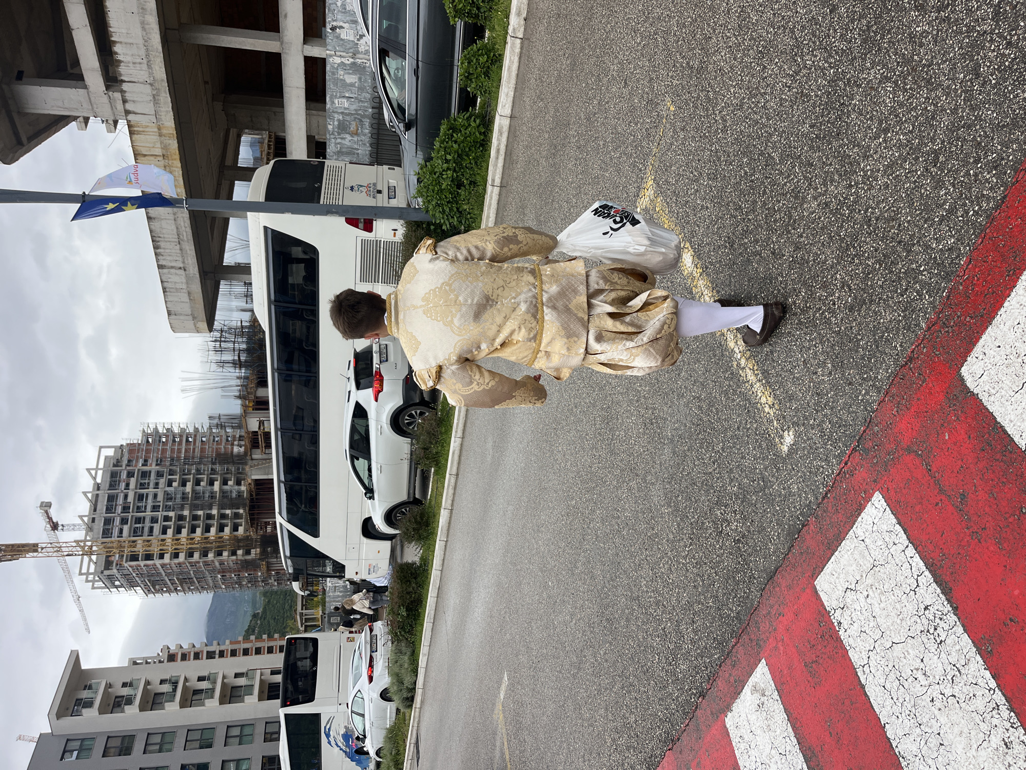A person dressed in an elaborate historical or theatrical costume is captured from behind, walking on an asphalt surface in an urban setting. The costume is rich gold and beige, featuring a patterned jacket and puffy breeches, paired with white stockings and dark, flat shoes. The individual carries a white plastic bag with a red and black logo (partially visible, possibly "OKTAN" or similar) in their left hand.
The ground is asphalt, marked with distinct red and white stripes, indicative of a crosswalk or a no-parking zone, and a yellow line near the person's feet. In the midground, several vehicles are parked, including a prominent white tour bus and other cars. A light pole stands tall, displaying two flags: one is the blue flag of the European Union with yellow stars, and the other is a white flag featuring the word "Budva" and a corresponding logo, confirming the location as Budva, Montenegro.
The background presents a dynamic cityscape. On the left, a large multi-story building is under active construction, with its concrete skeleton and scaffolding clearly visible, alongside a construction crane. Other completed modern buildings are also present. Distant mountains frame the scene under an overcast sky. The combination of historical attire with modern elements like the plastic bag, construction, and tour buses creates an interesting juxtaposition, possibly indicating a local event, festival, or performance taking place in Budva.