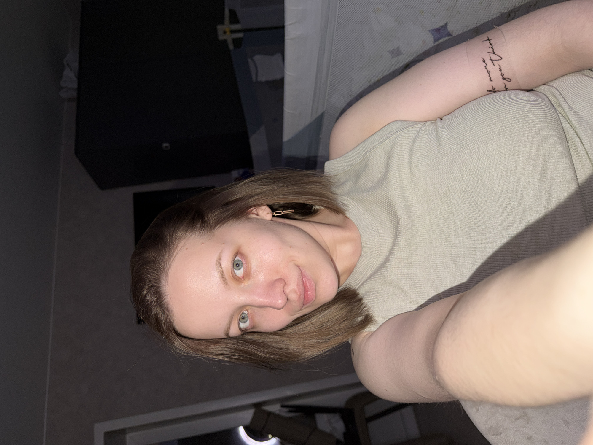 The image, rotated 90 degrees clockwise, features a young woman taking a selfie indoors. She is positioned towards the lower left of the frame, looking directly at the camera with a neutral to slight smile.
The woman has fair skin, short to medium-length brown hair, and striking blue-green eyes. A small, golden rectangular hair clip is visible on the right side of her head (viewer's left). She is wearing a light beige or pale green ribbed high-neck top. On her right forearm (viewer's left arm), there is a tattoo written in a delicate script font that reads "be more of you."
A strong, warm light source illuminates her face and arm from the upper right, casting diagonal shadows across her body and the background. The background is largely dark and indistinct, with a large, dark rectangular object (possibly a television or monitor) situated behind her head. To the upper right, a lighter, textured surface, possibly a bed frame or furniture, is visible. The setting appears to be a casual indoor space, likely a bedroom, located in Reshetnikovo, Russia. The lighting suggests it is daytime, with some areas of the room appearing shadowed.