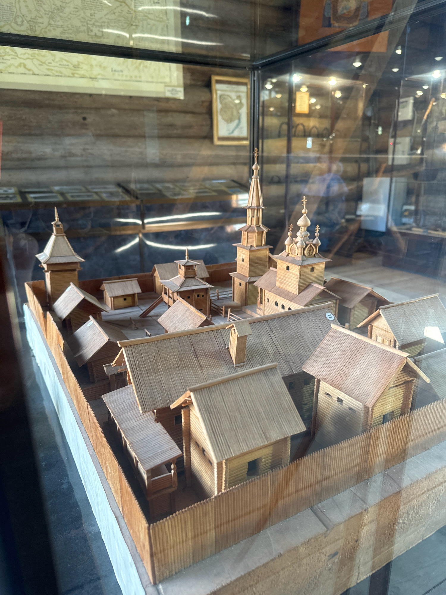An indoor, eye-level, medium shot captures a detailed wooden diorama of a fortified settlement inside a glass display case. The diorama depicts a cluster of wooden buildings, including a church with multiple onion domes and spires, and a tall watchtower, all enclosed by a palisade fence. The buildings are constructed with meticulously crafted miniature wooden logs and planks, featuring distinct roofs, doorways, and windows. Inside the display case, behind the diorama, shelves hold other museum artifacts, some of which are indistinct due to reflections on the glass. A framed map is visible on the wall above the diorama, and other framed items are displayed on the shelves in the background. The diorama is positioned on a wooden base. The overall setting appears to be a museum in the Taltsy (Irkutsk) Museum of Wooden Architecture and Ethnography, Russia, showcasing historical architecture.